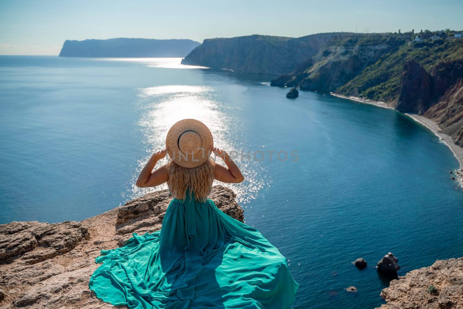 Woman sea. A happy girl is sitting with her back to the viewer in a mint dress on top of a mountain against the background of the ocean and rocks in the sea