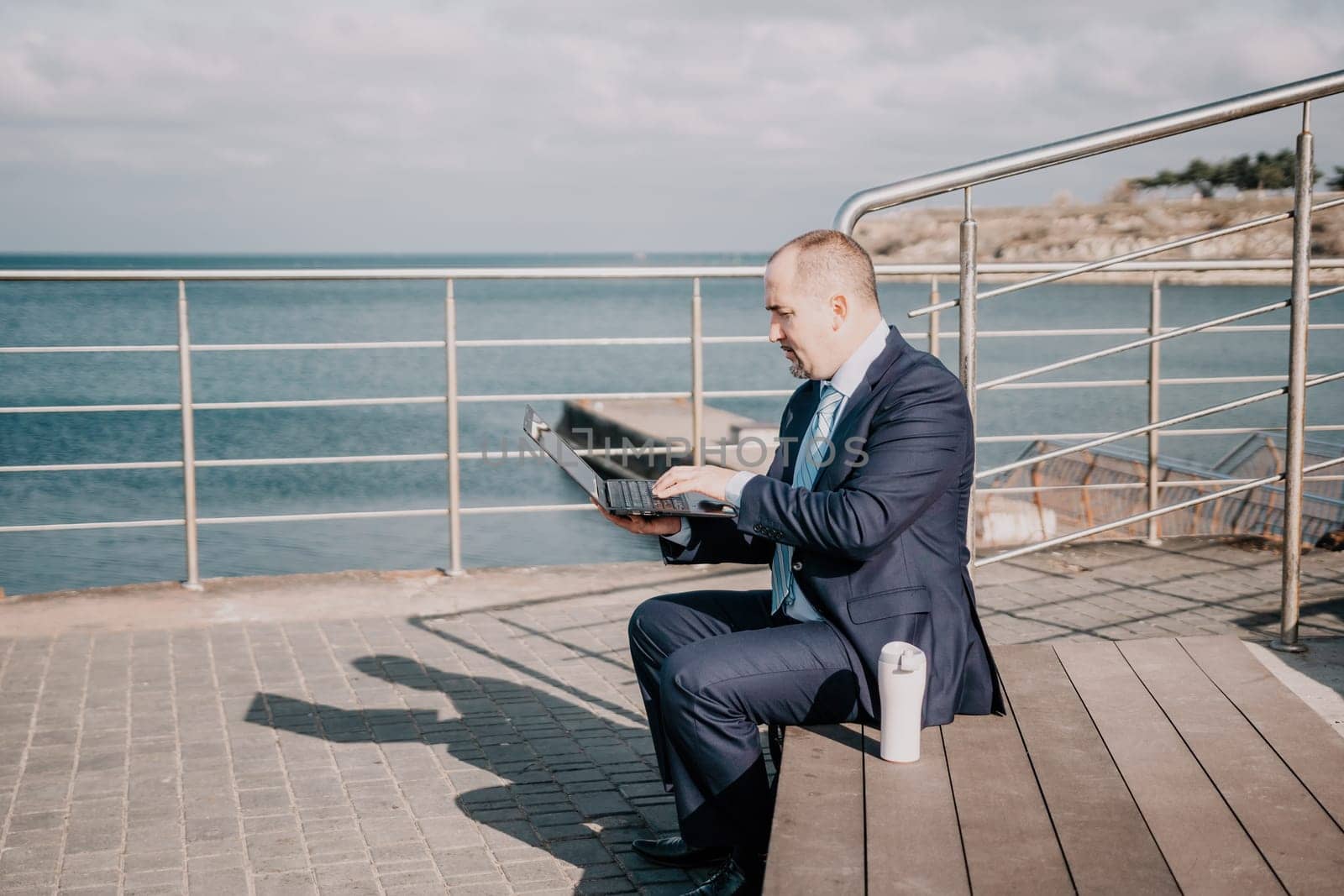 Confident middle age businessman working remotely online, typing on a laptop keyboard while sitting on a beach at sunset. Working remotely on vacation, running an online business from a distance. by panophotograph