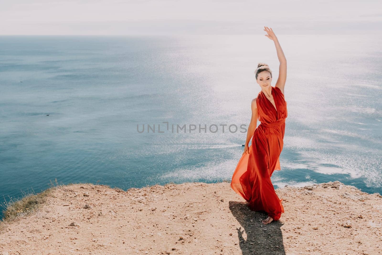 Side view a Young beautiful sensual woman in a red long dress posing on a rock high above the sea during sunrise. Girl on the nature on blue sky background. Fashion photo.