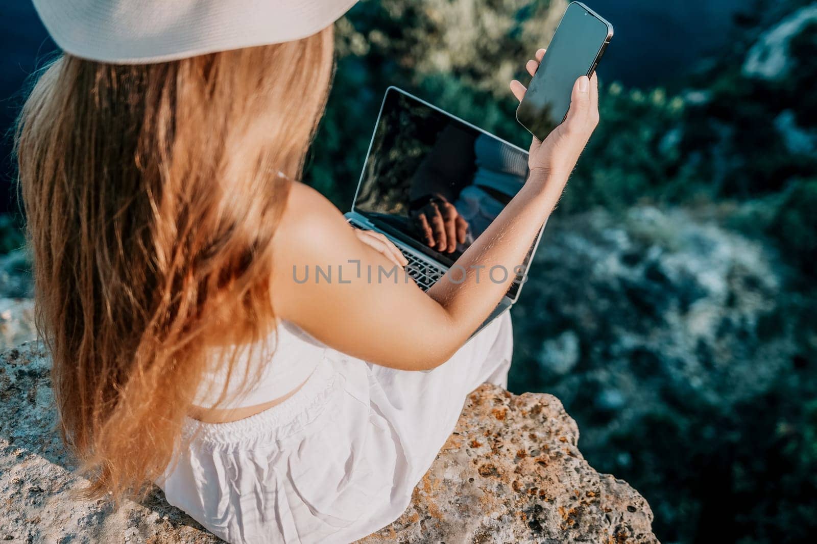 Successful business woman in yellow hat working on laptop by the sea. Pretty lady typing on computer at summer day outdoors. Freelance, travel and holidays concept.