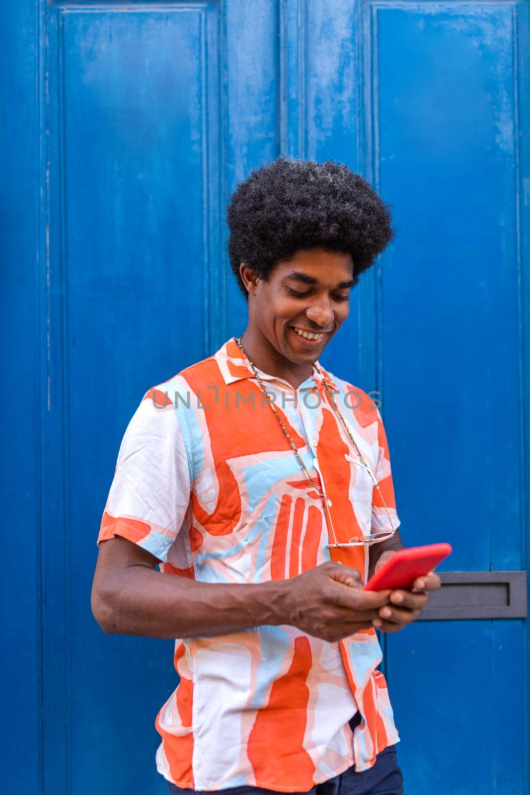 African American man using mobile phone to text a friend standing in the street. Blue door background. Vertical image. Colourful image.