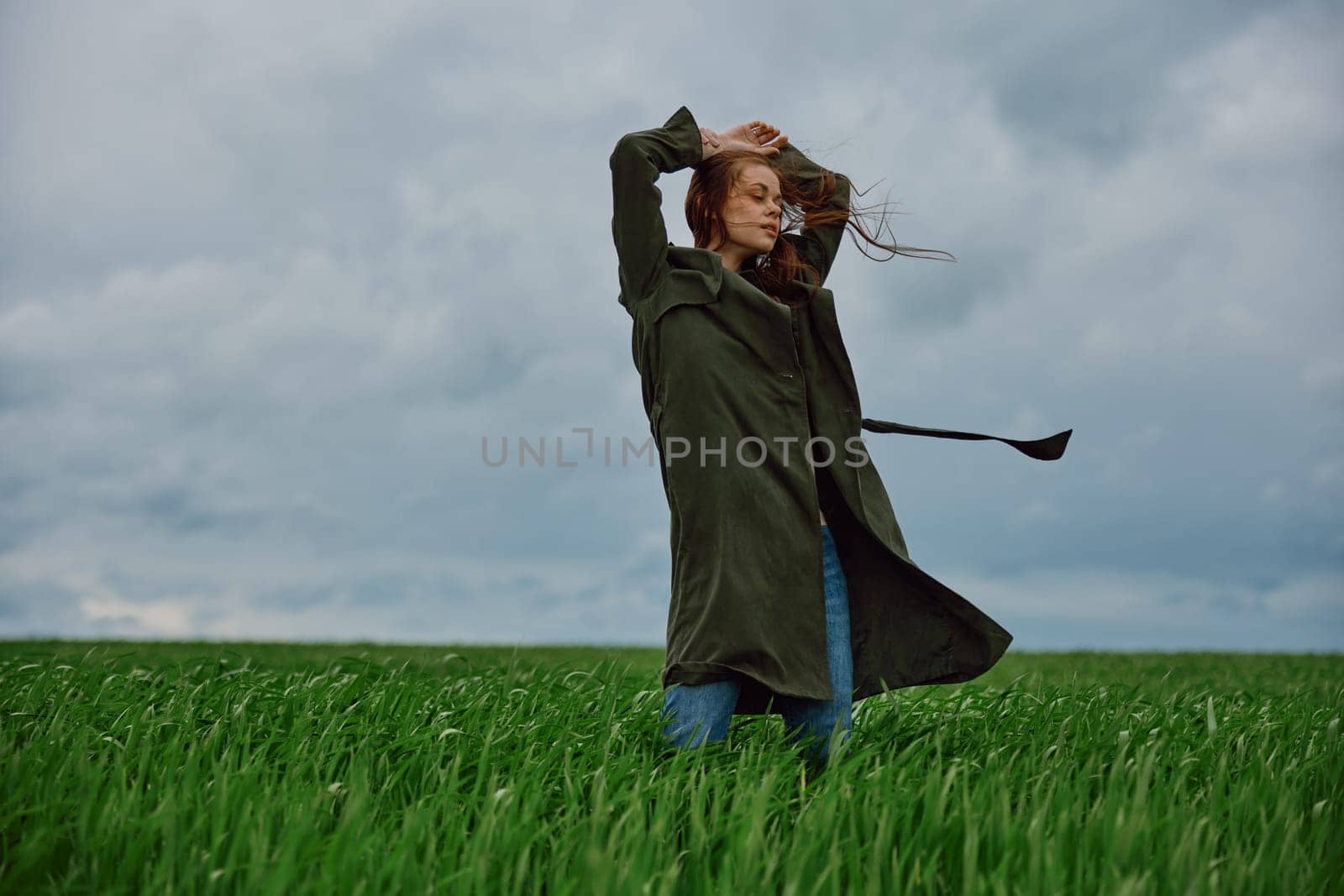a woman in a long coat stands in a field against a cloudy sky with her back to the wind with her hands raised behind her head. High quality photo
