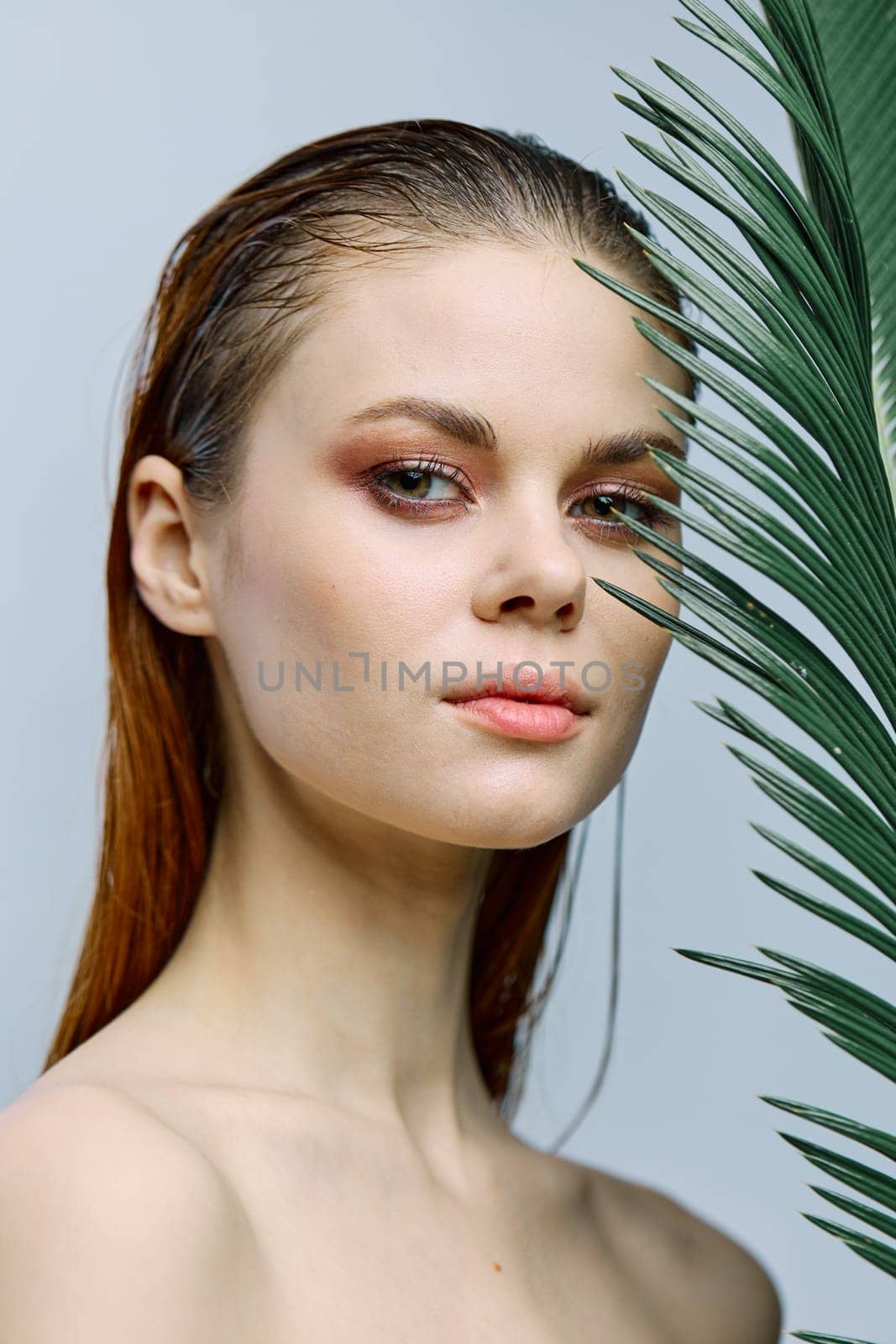 a close beauty portrait of a beautiful woman standing on a light background holding a palm leaf near her face, looking into the camera. Vertical photo without retouching of problem skin. High quality photo