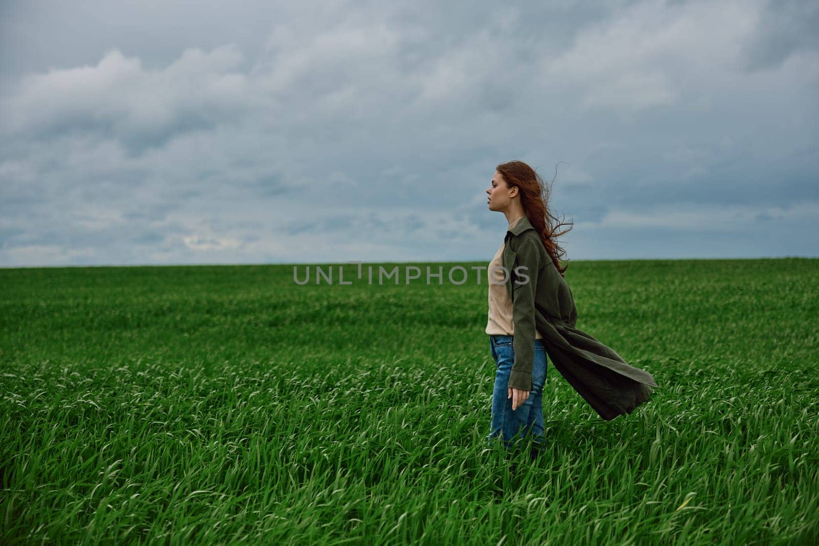 a beautiful woman stands in a green field in a raincoat and looks into the distance. Strong wind, flying hair, harmony with nature by Vichizh