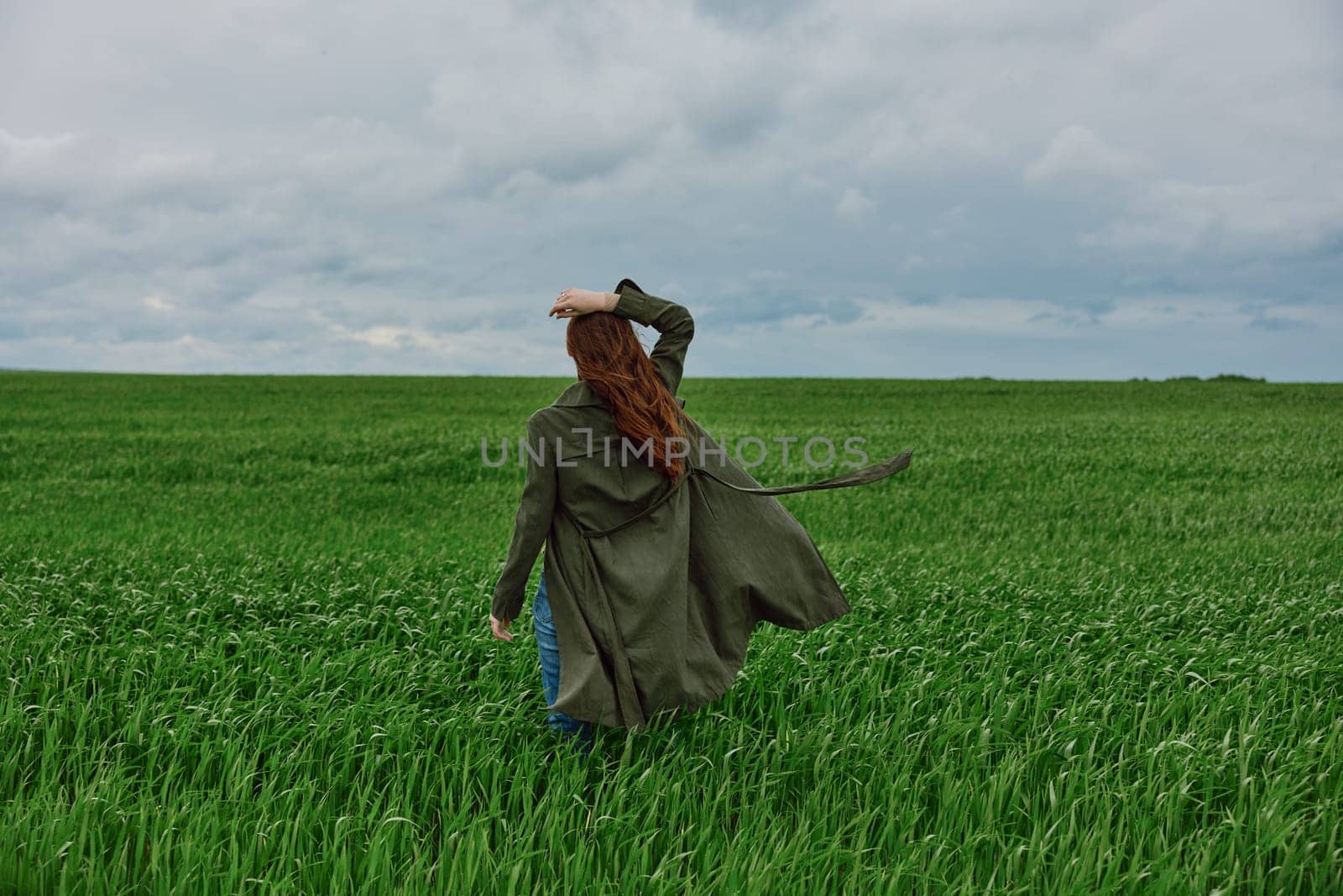 a red-haired woman stands with her back to the camera in a long coat, holding her hand with her hair flying in the wind, standing in a green field in cloudy weather. High quality photo