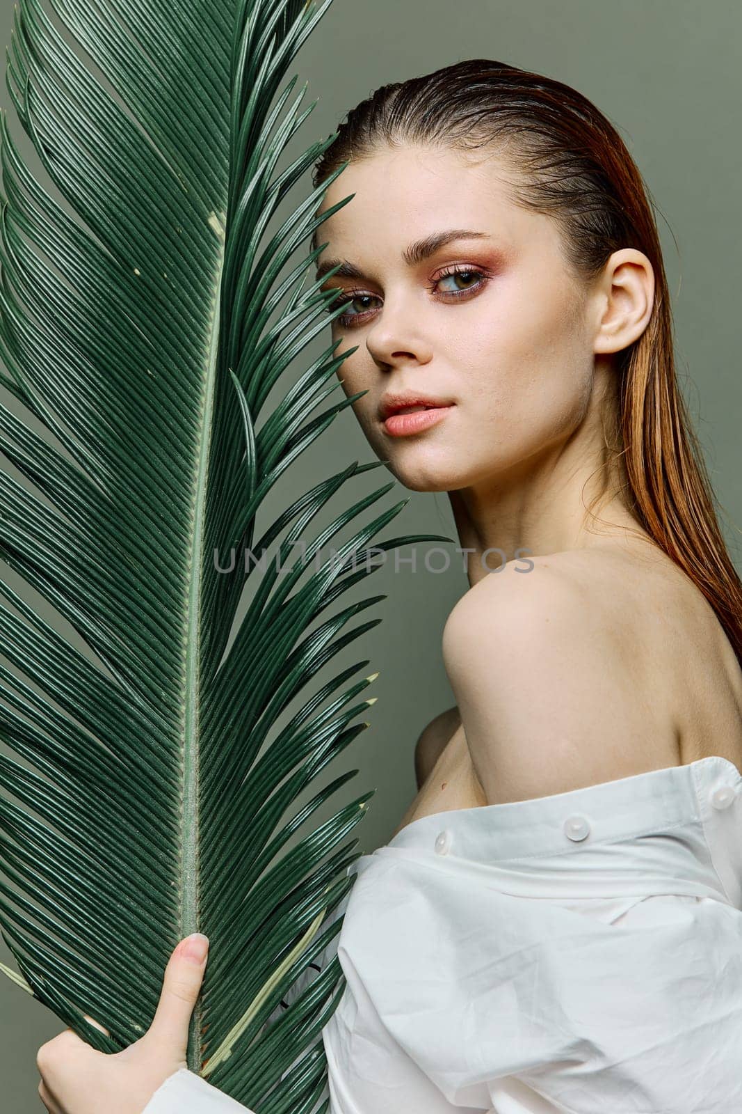 a sophisticated, elegant woman with evening makeup, stands with a green palm leaf, dressed in a light shirt. Close vertical portrait, photo without retouching. High quality photo