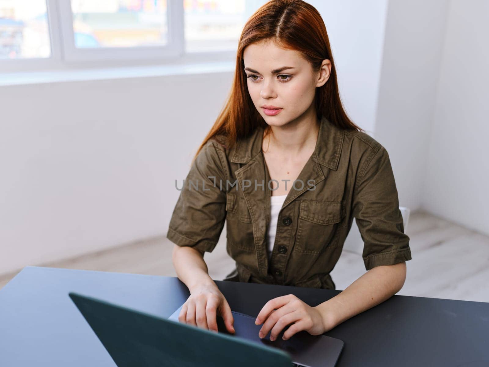 a woman sits at a table in a bright office with a laptop against the background of a window by Vichizh