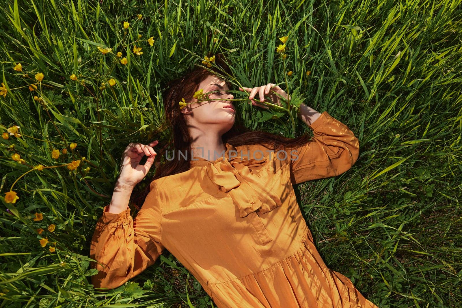 a calm woman with long red hair lies in a green field with yellow flowers, in an orange dress with her eyes closed, spreading her arms to the sides, enjoying peace and recuperating. High quality photo