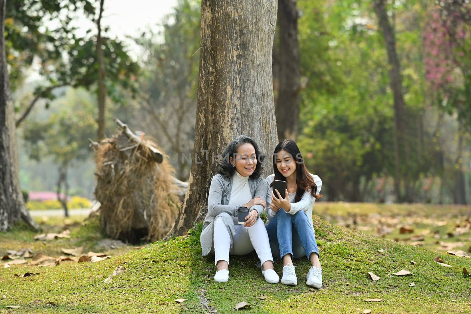 Photo of smiling senior mother and adult daughter using smart phone while sitting on green grass against calm nature park by prathanchorruangsak