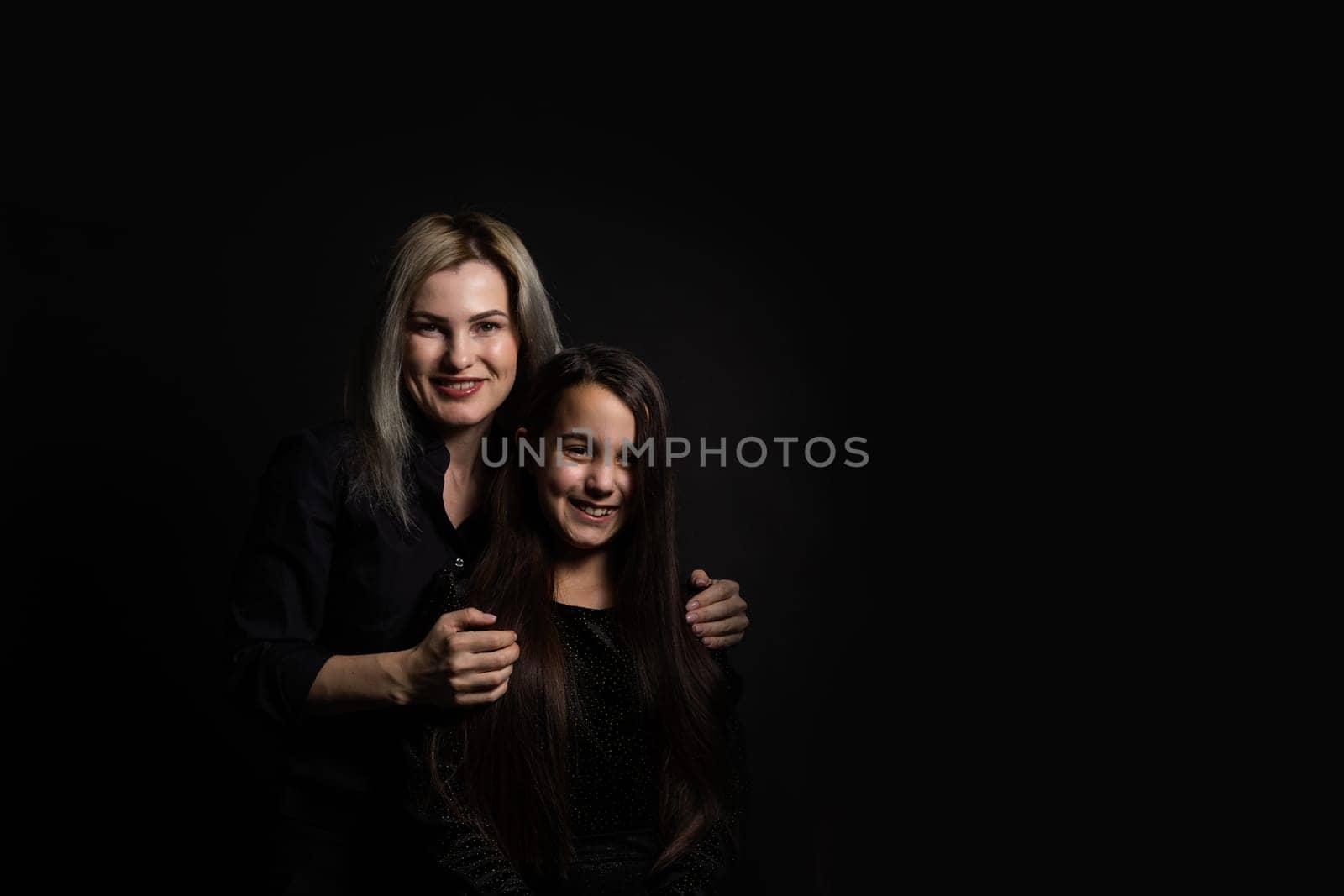 Young mother and her little daughter are looking at camera and smiling, sitting against black wall.