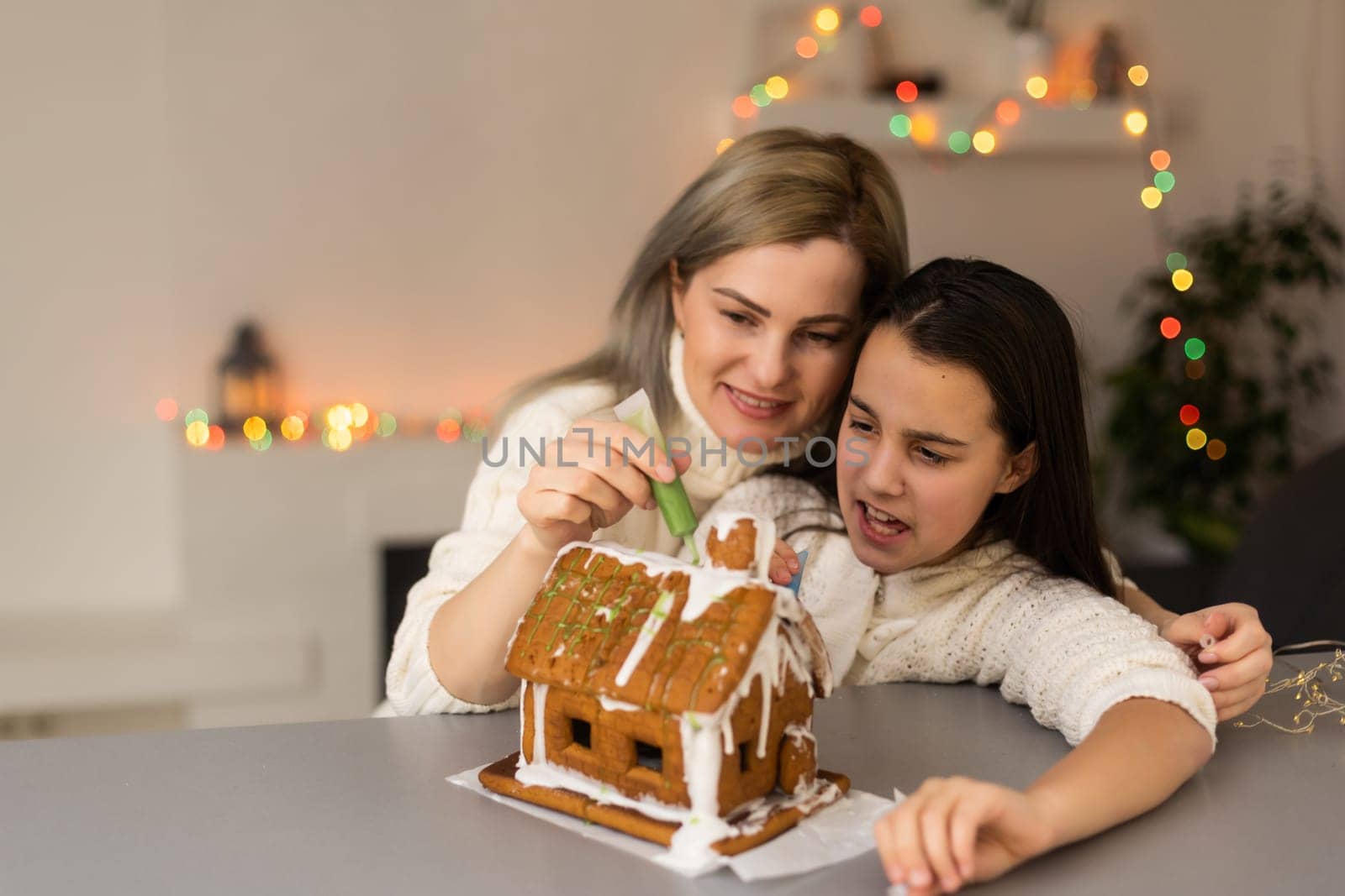 mother and daughter decorating gingerbread house. Beautiful living room with lights. Happy family celebrating holiday together