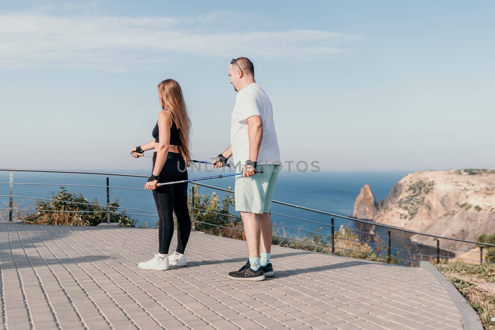 Happy Middle aged couple or friends practicing nordic walking in park near sea. Mature couple with trekking poles walking, practicing Nordic walking outdoors. Aging youthfully and sport concept by panophotograph