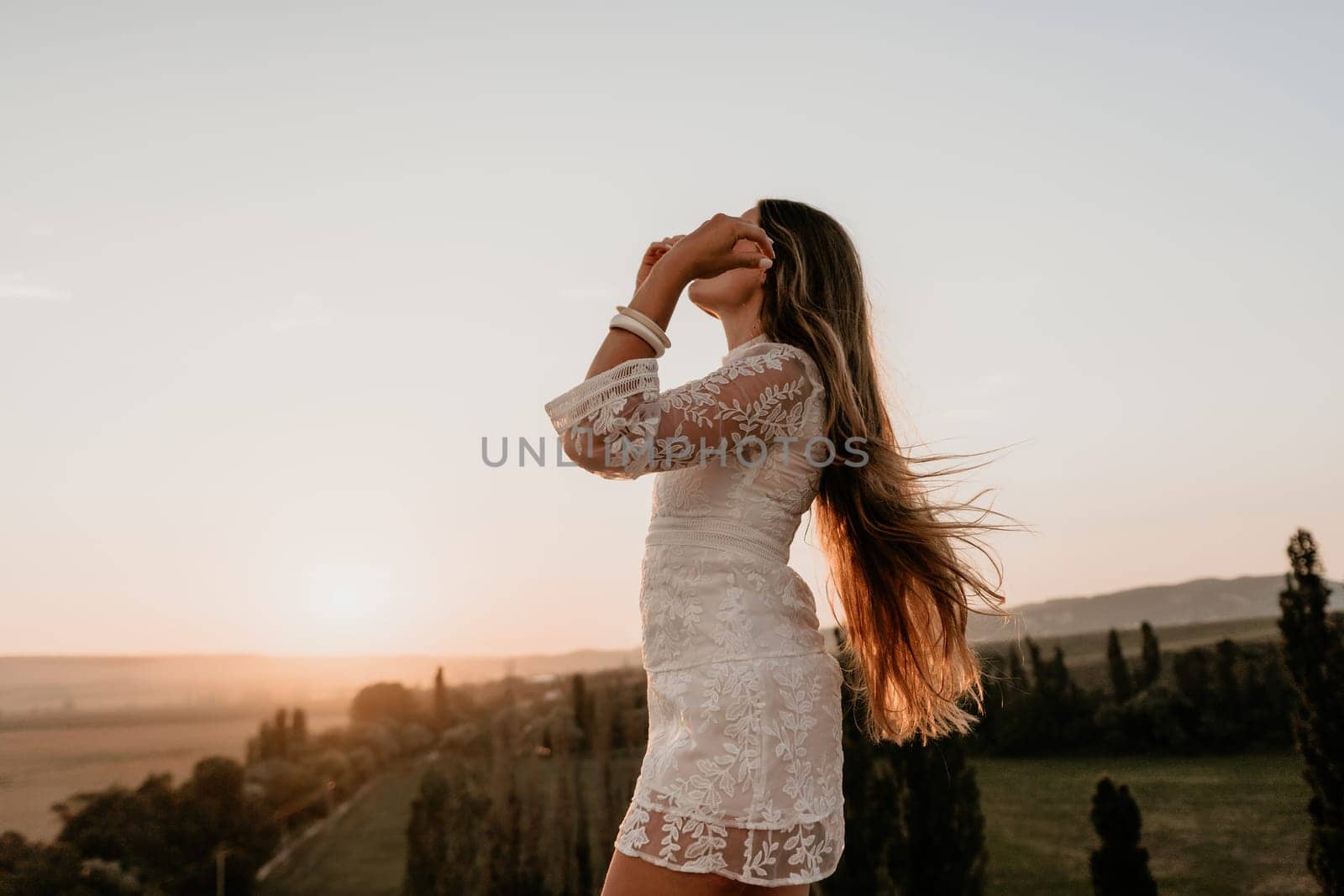 Happy woman in white boho dress on sunset in mountains. Romantic woman with long hair standing with her back on the sunset in nature in summer with open hands. Silhouette. Nature. Sunset. by panophotograph