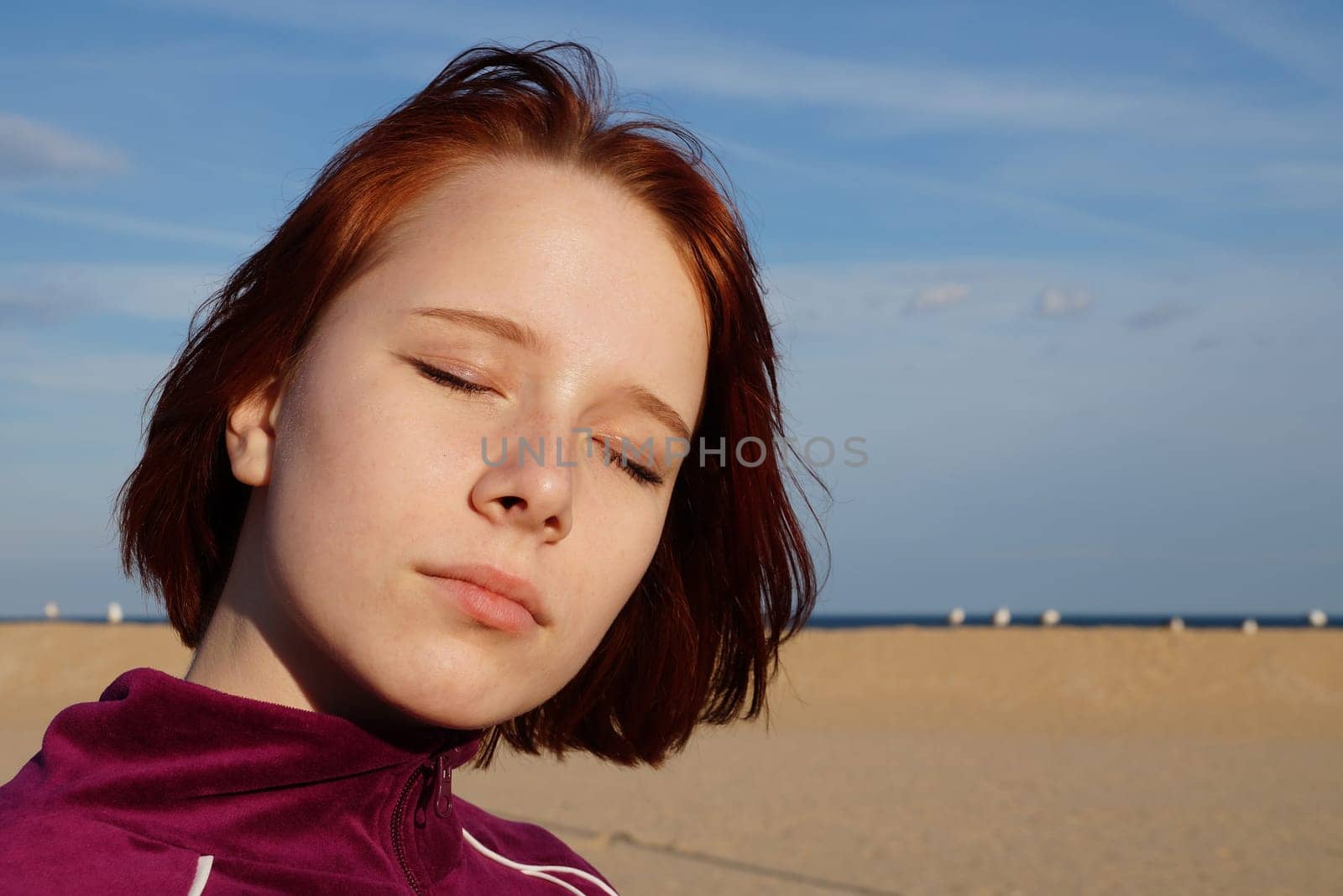 Portrait of a red-haired teenage girl with closed eyes on an empty beach.