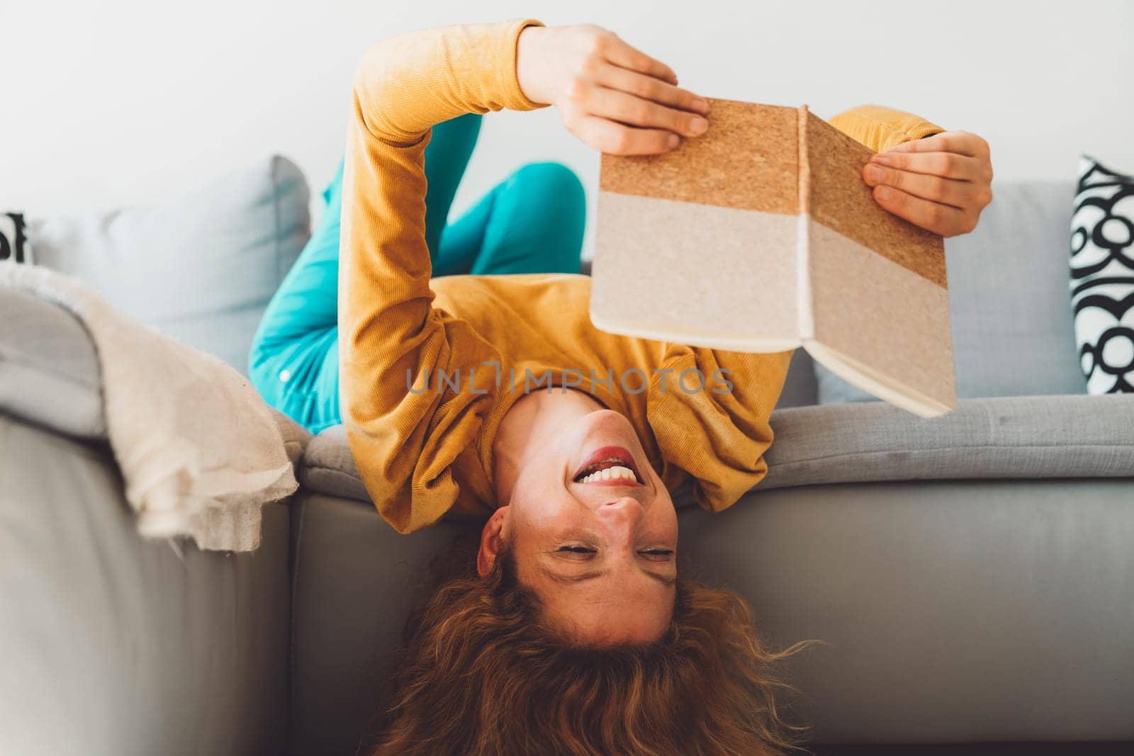 Cheerful woman laying on the couch with her head hanging down while holding a notebook in her hands by VisualProductions