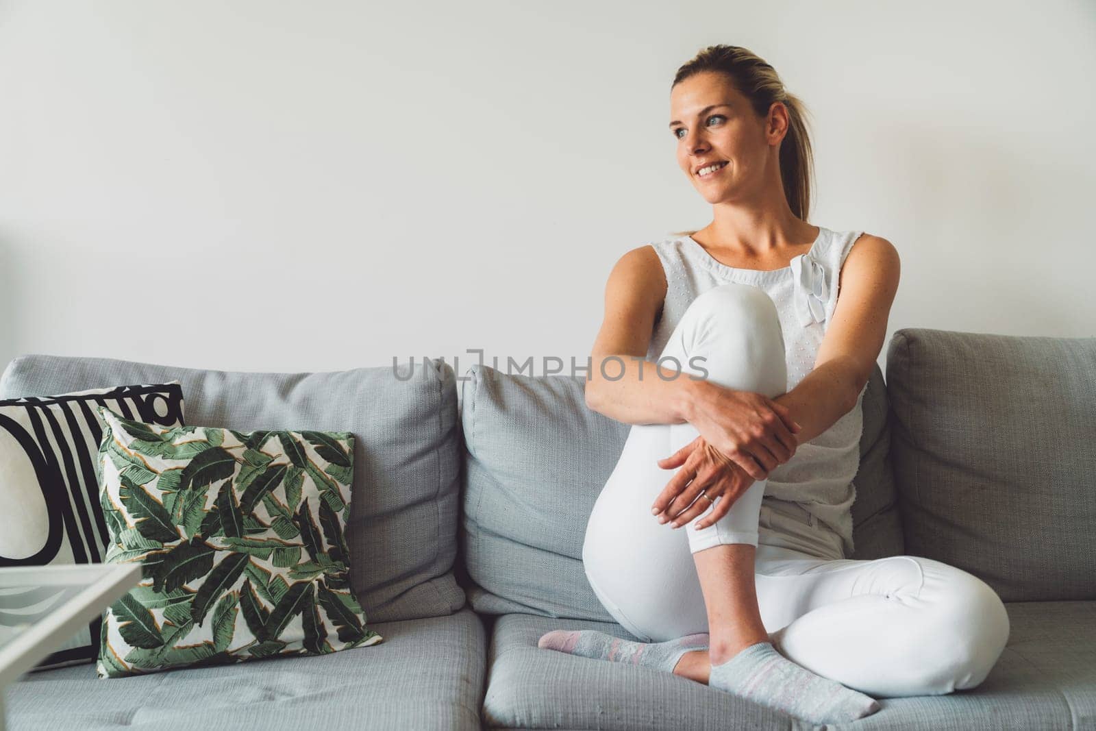 Cheerful smiling woman relaxing on the couch at home, dressed in white jeans and white shirt by VisualProductions
