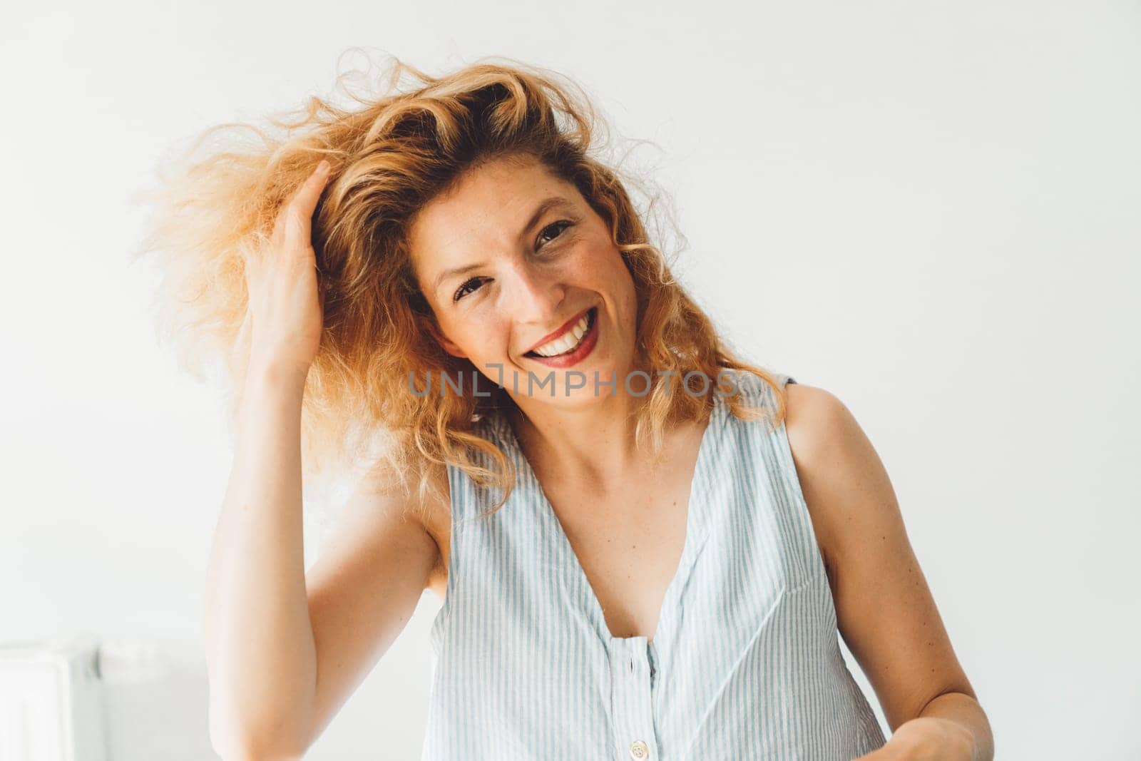 Waist up portrait smiling cheerful woman with curly hair and red lipstick looking at the camera, white wall background by VisualProductions