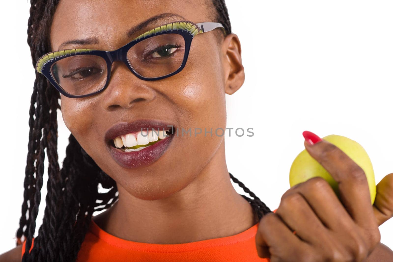 young girl standing in glasses on white background eating an apple while looking at the camera.