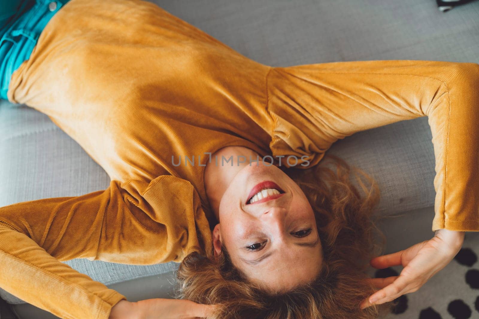 Smiling woman laying on the couch in a velvet yellow shirt playing with her hair looking at the camera by VisualProductions