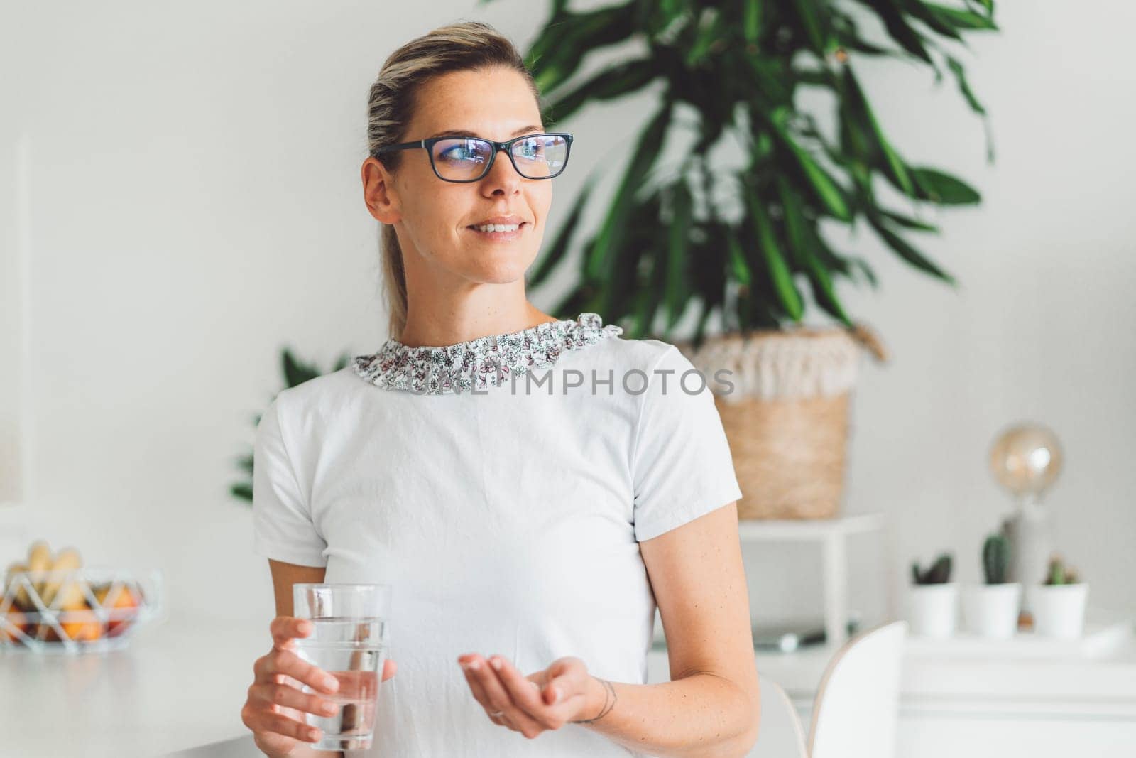 Smiling young caucasian woman taking her morning supplements, vitamin pills with a glass of water at home. High quality photo