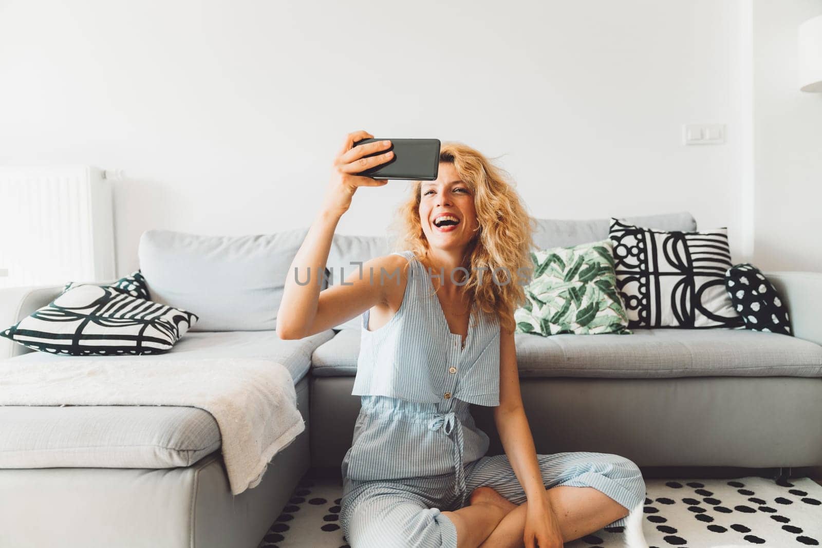 Cheerful curly blonde woman sitting on the carpet in front of the sofa taking selfies by VisualProductions
