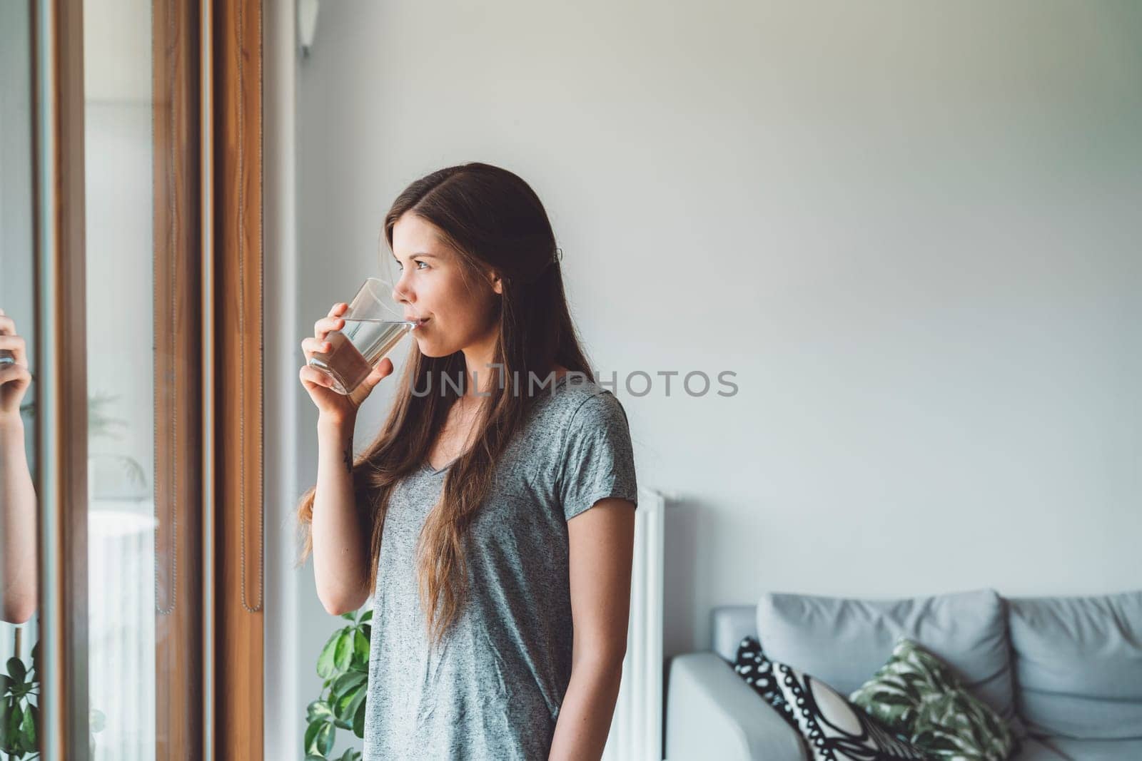 Young woman with long brown hair standing by the window looking out while drinking a glass of water and taking her vitamin pills by VisualProductions