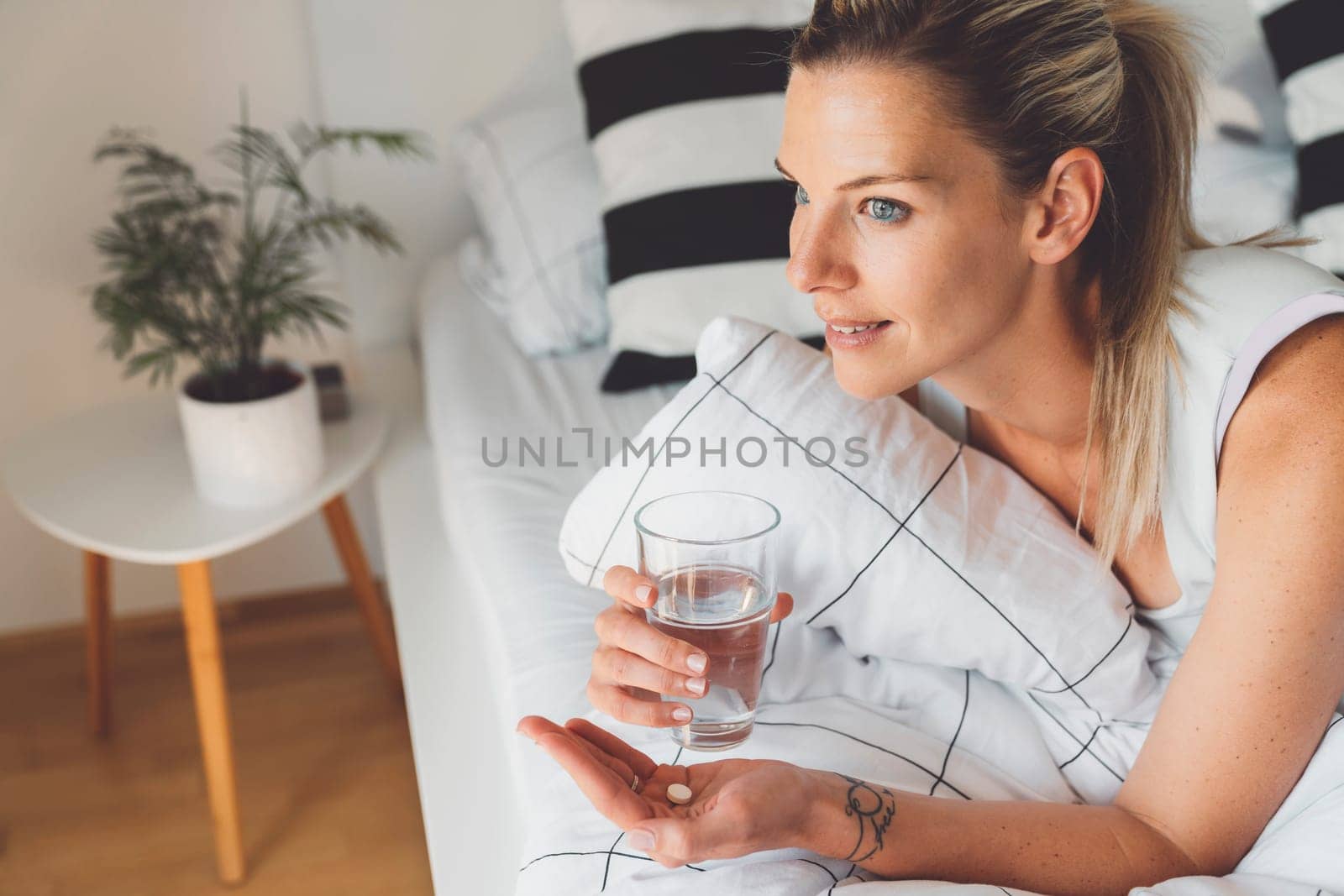 Close up portrait young woman still in bed in the morning holding a glass of water in one hands and vitamin pill in the other by VisualProductions