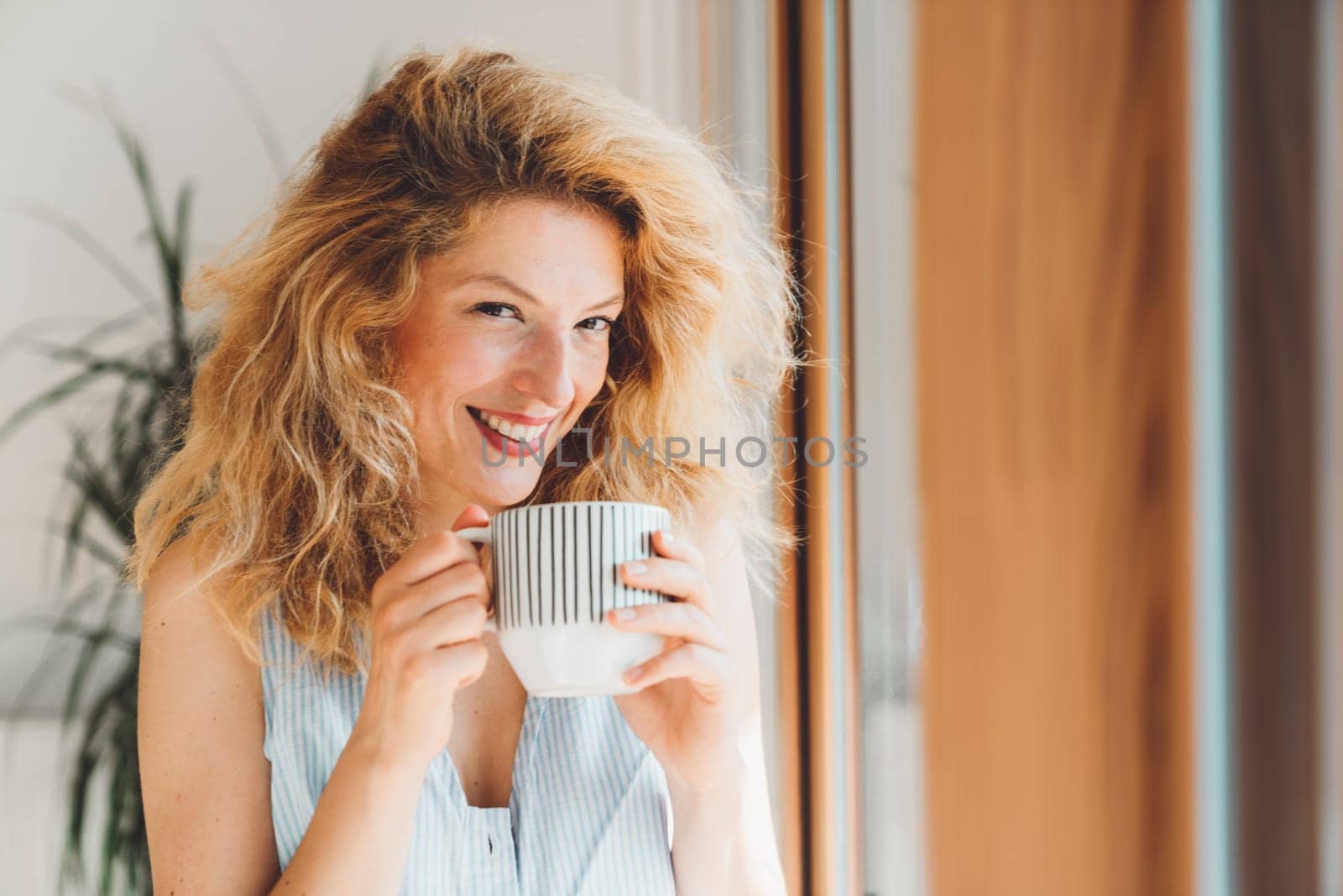 Waist up portrait of a smiling woman with curly har holding a cup of tea standing by the window looking at the camera by VisualProductions
