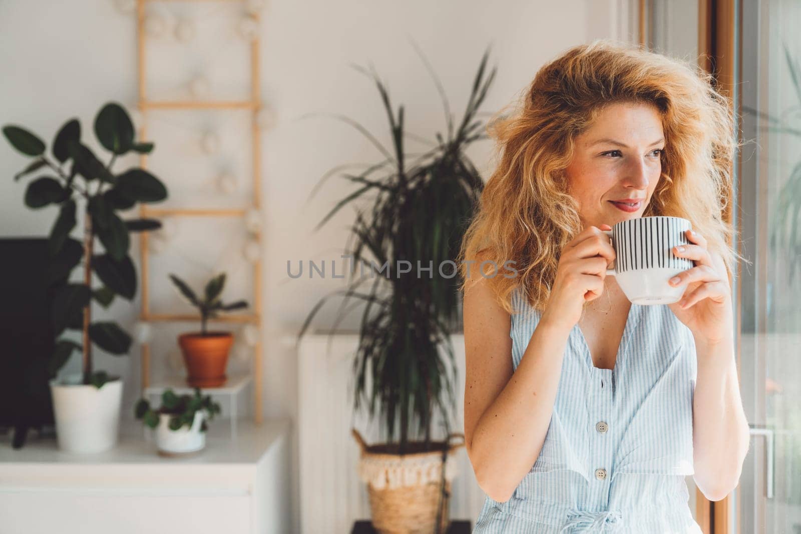 Waist up portrait woman with blonde curly hair holding a cup of tea standing by the window looking out, plants in the background by VisualProductions