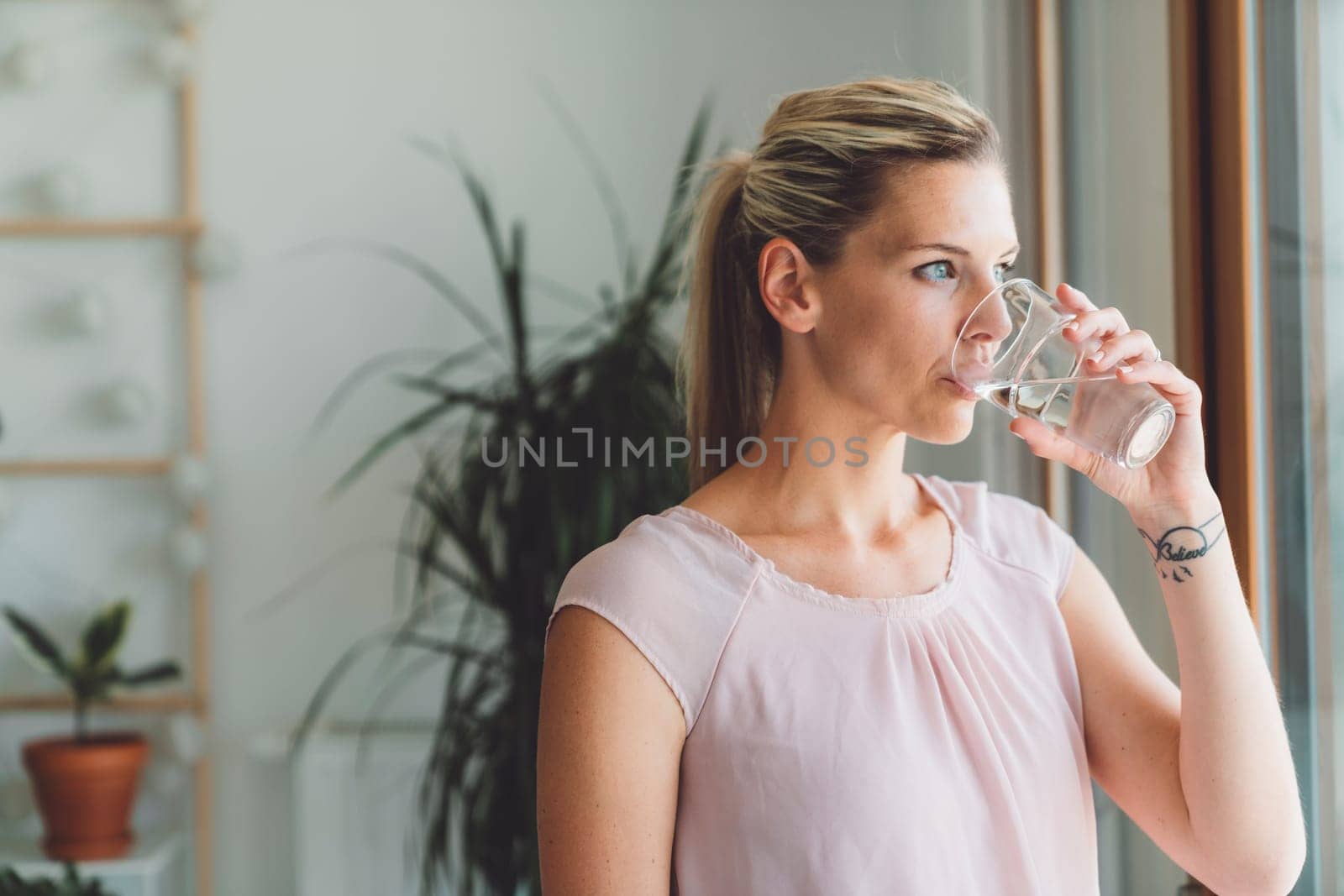 Portrait of blonde woman with a pony tail drinking a glass of water while standing by the window of her apartment by VisualProductions