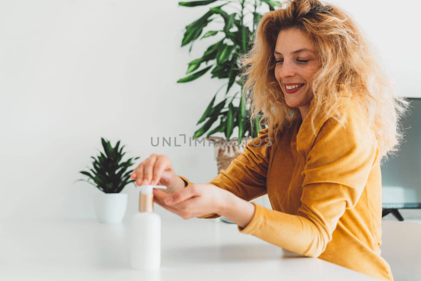 Smiling curly woman wait up sitting by the table using her hand cream by VisualProductions