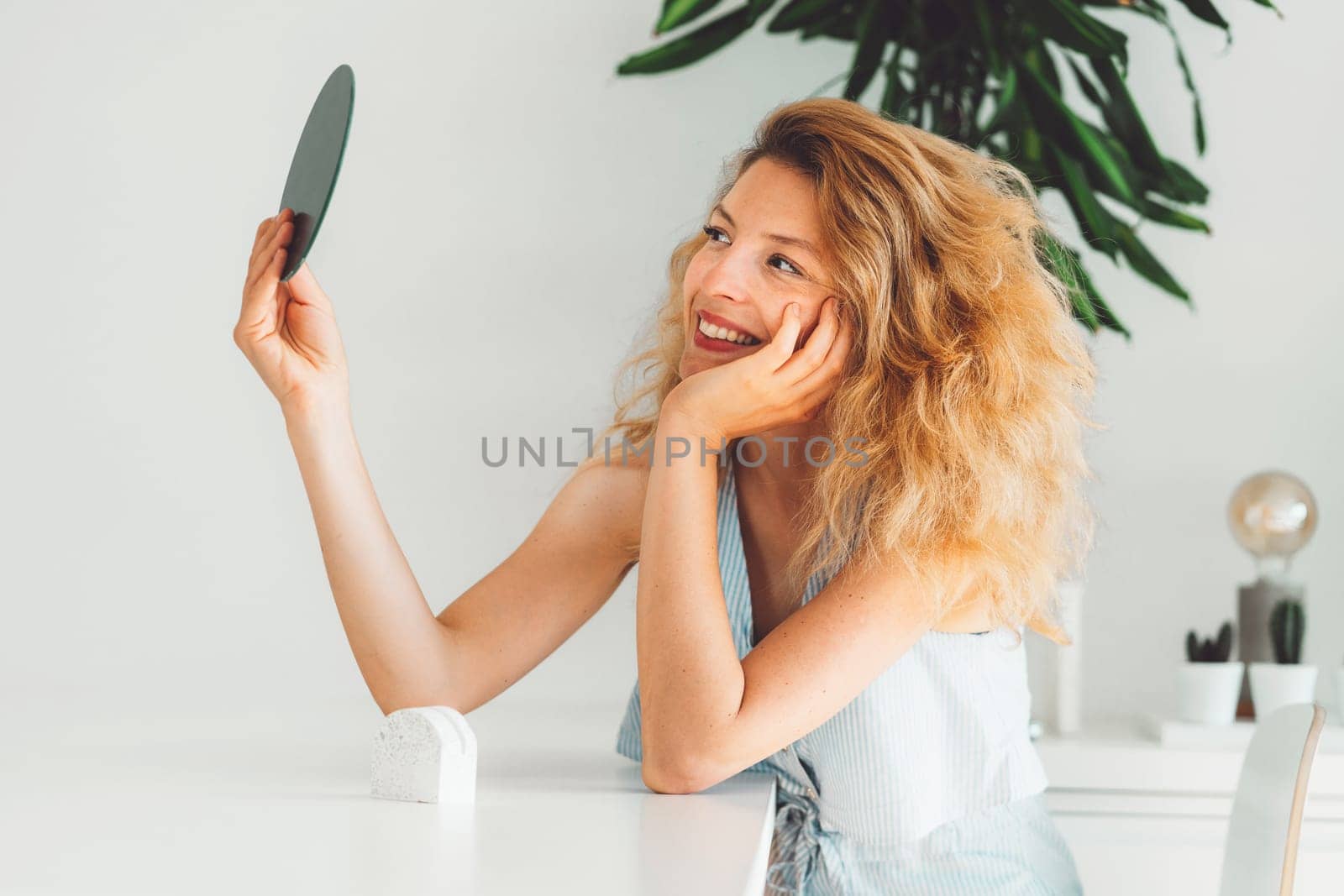 Smiling blonde woman with curly hair looking at herself in a small round mirror she is holding up by VisualProductions