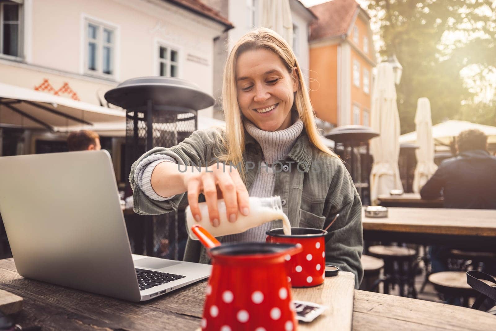 Smiling blonde woman pouring milk in her coffee sitting outside at a cafe, while working on her laptop by VisualProductions