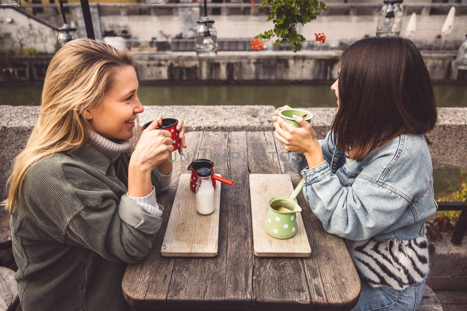 Two young women blonde and brunette sitting at a cafe by the river having coffee and a talk by VisualProductions