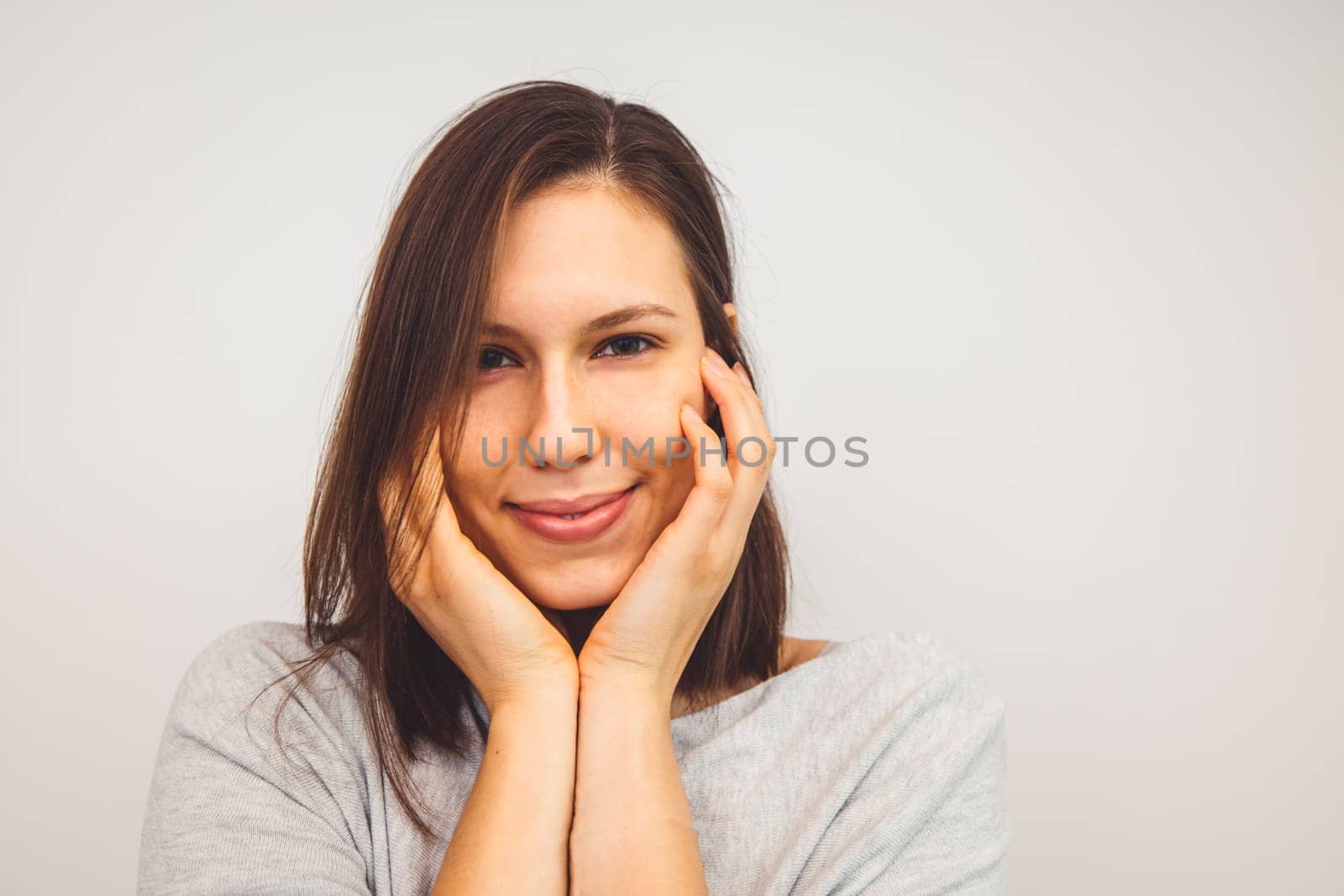 Portrait of young woman holding hands on her face with a sbtle smile looking at the camera on white background by VisualProductions