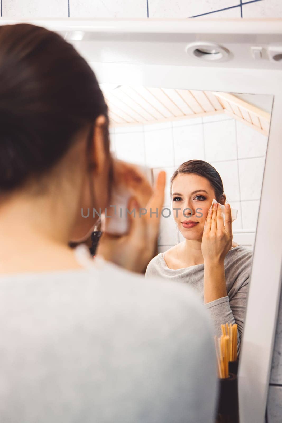 Over the shoulder view, young woman cleaning make up off her face standing in front of a mirror by VisualProductions