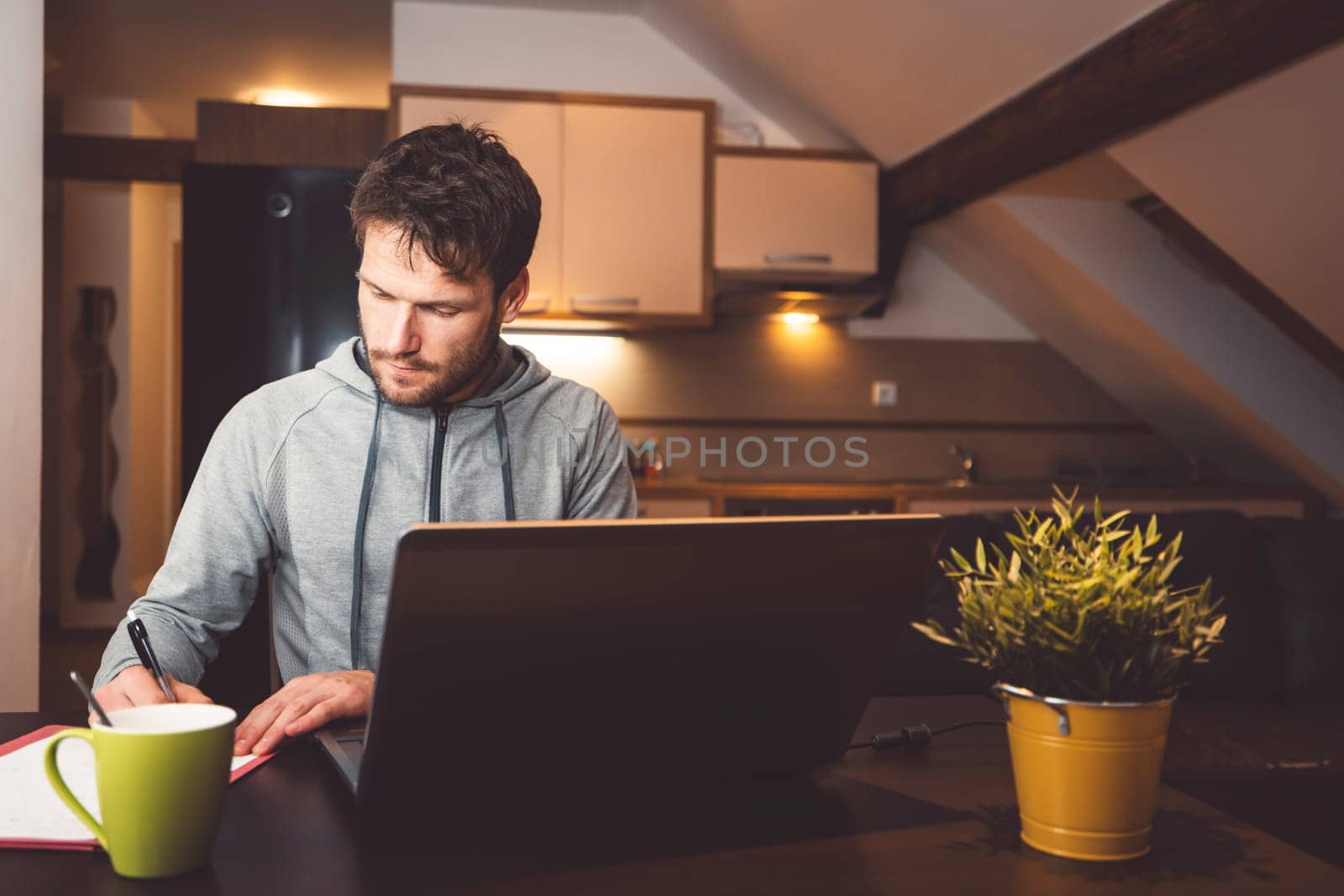 Caucasian man writing notes while attending evening online studying lessons on his laptop, sitting by the table with a cup of coffee next to the laptop by VisualProductions