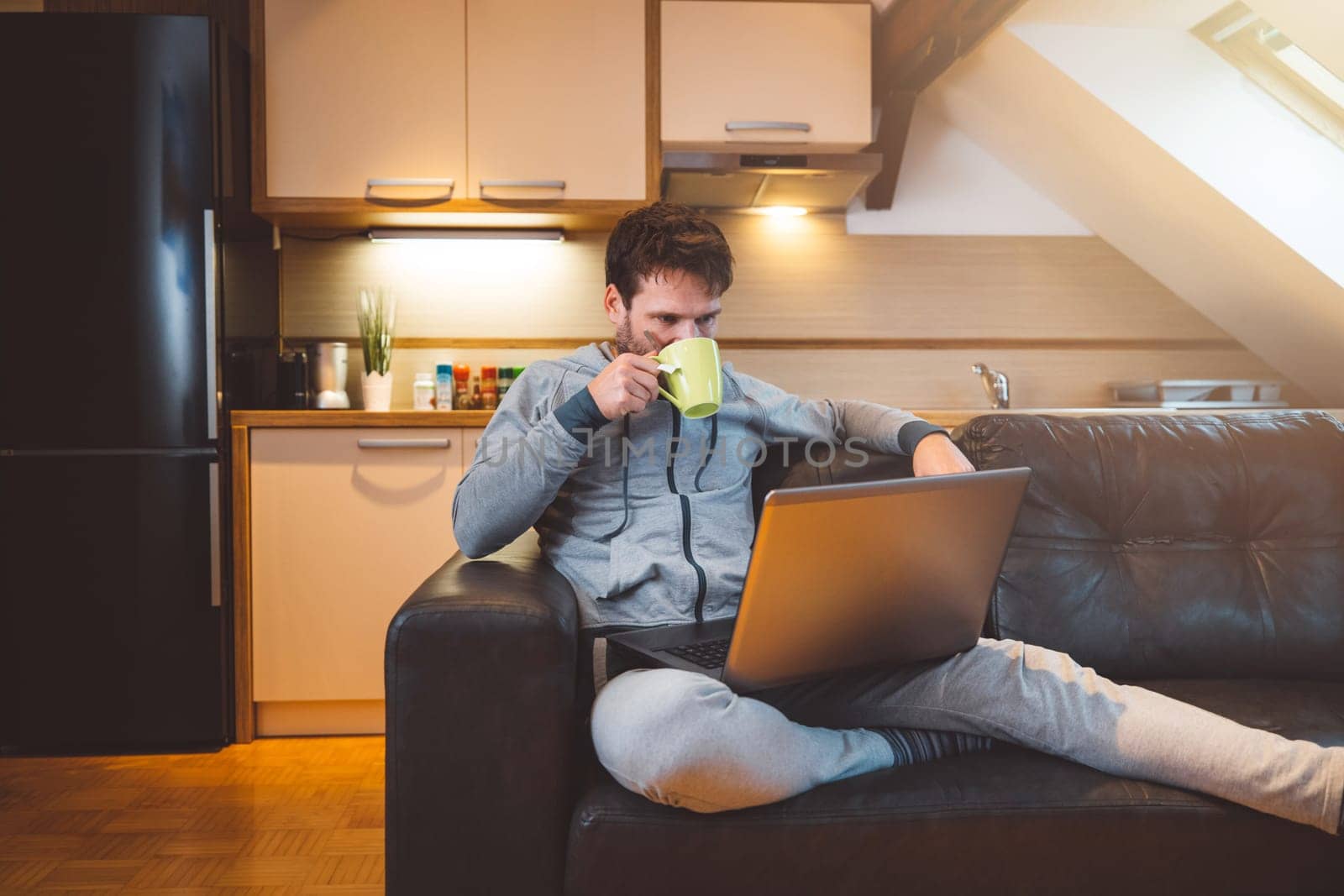 Man drinking coffee while laying on the couch with laptop on his lap, doing online courses by VisualProductions