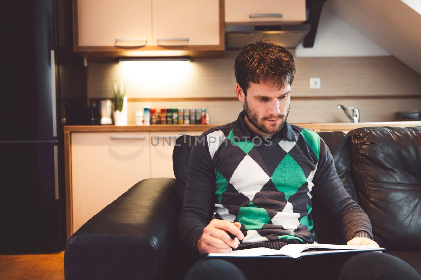Waist up adult man in green and black sweater sitting on a leather sofa studying for exam at home by VisualProductions