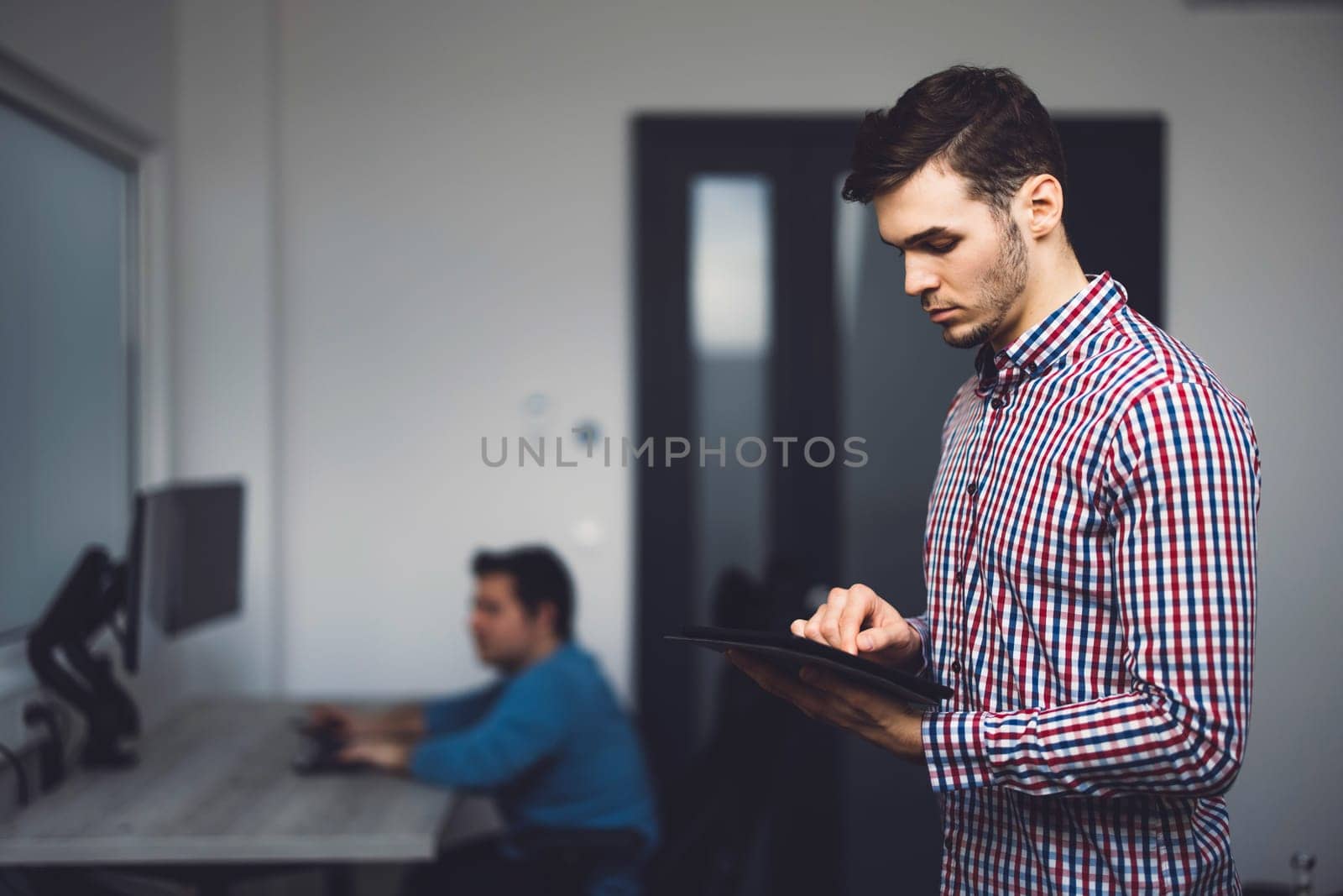 Side view of young programmer standing in the office looking down at the digital tablet he is holding in his hands by VisualProductions