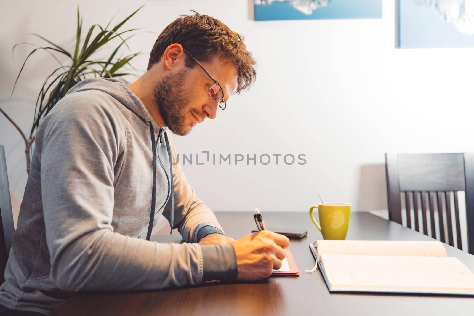 Side view of man with glasses wearing a tracksuit, sitting by the table making notes studying at home by VisualProductions