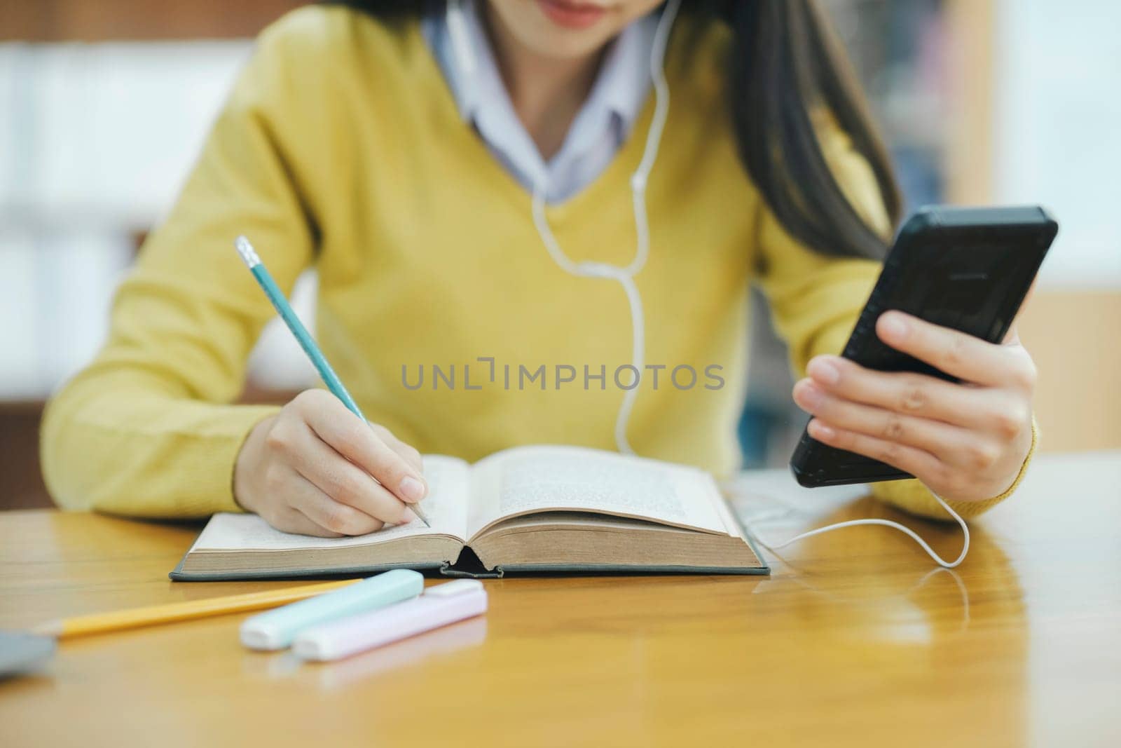 Young female college highschool student with headphones and in casual cloths sitting at desk studying, reading books, and writing with handphone at library for research or school project. E-Learning Education Library concept.