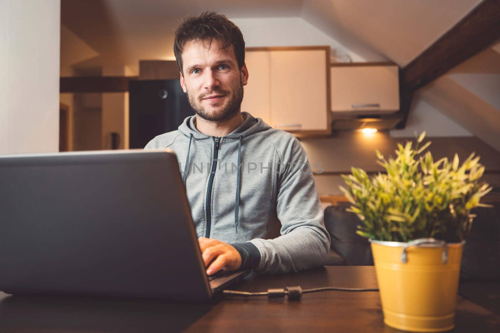 Front view of a man sitting by the table working on his laptop from home in the evening, looking at the camera by VisualProductions