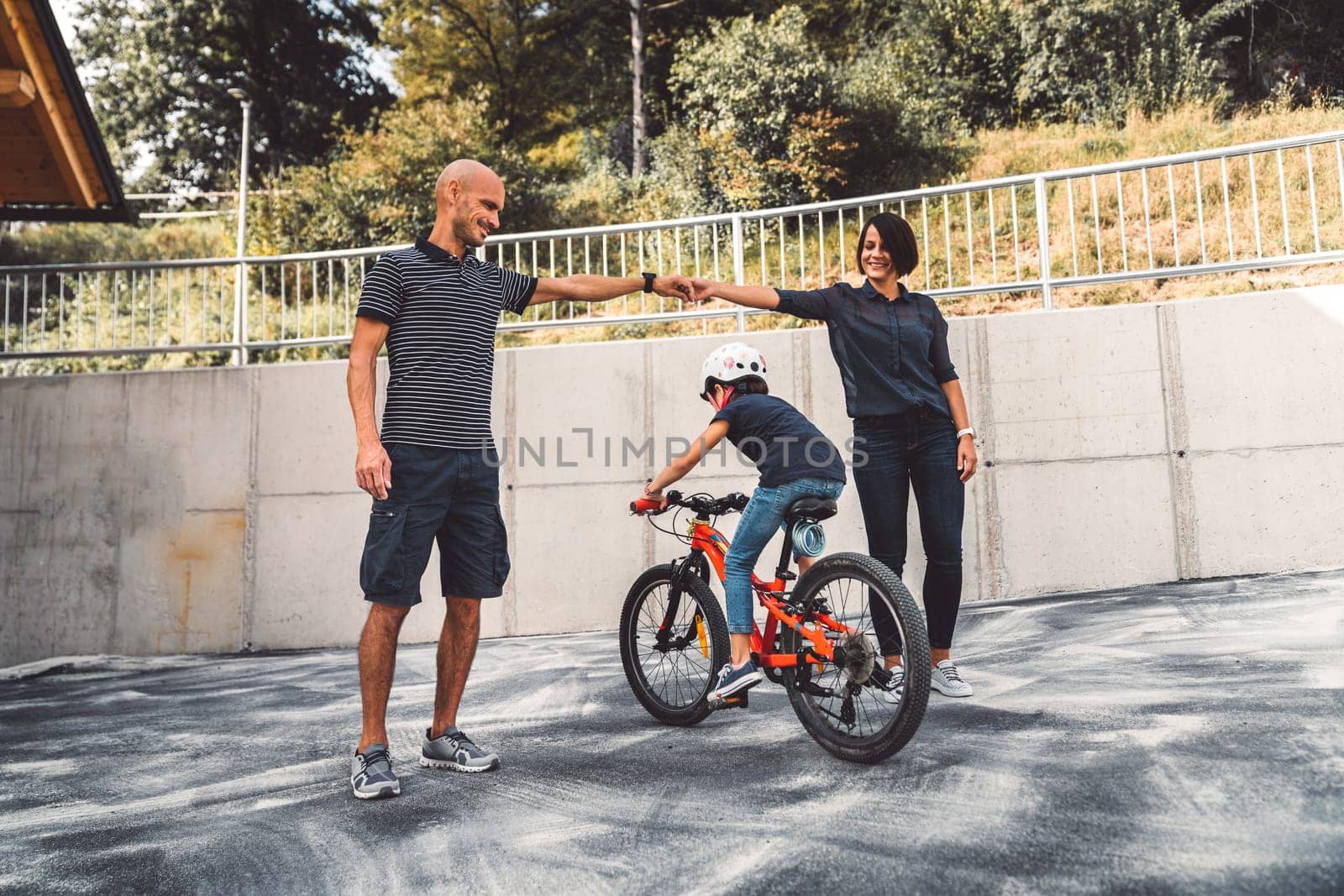 Family of three having fun outside, girl riding a bike, while parents hold hands for their daughter to ride underneath by VisualProductions