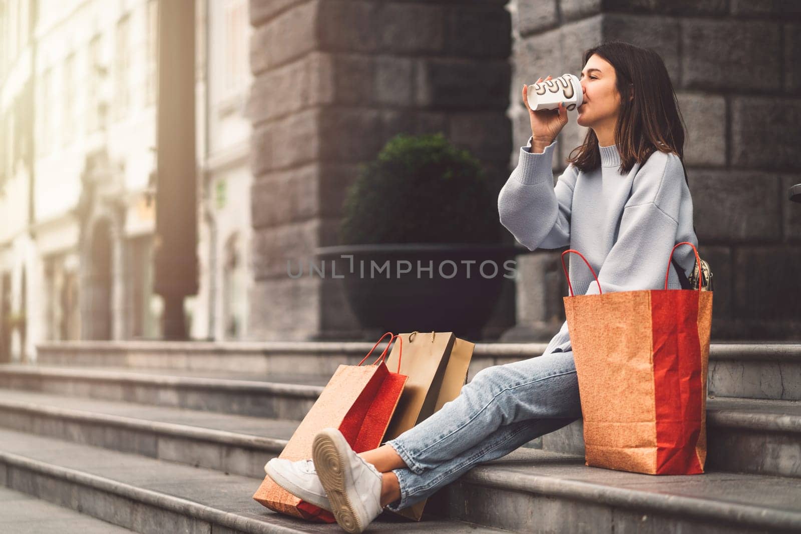 Young woman sitting on the stairs in the city, drinking coffee from a to go cup, with paper shopping bags around her by VisualProductions