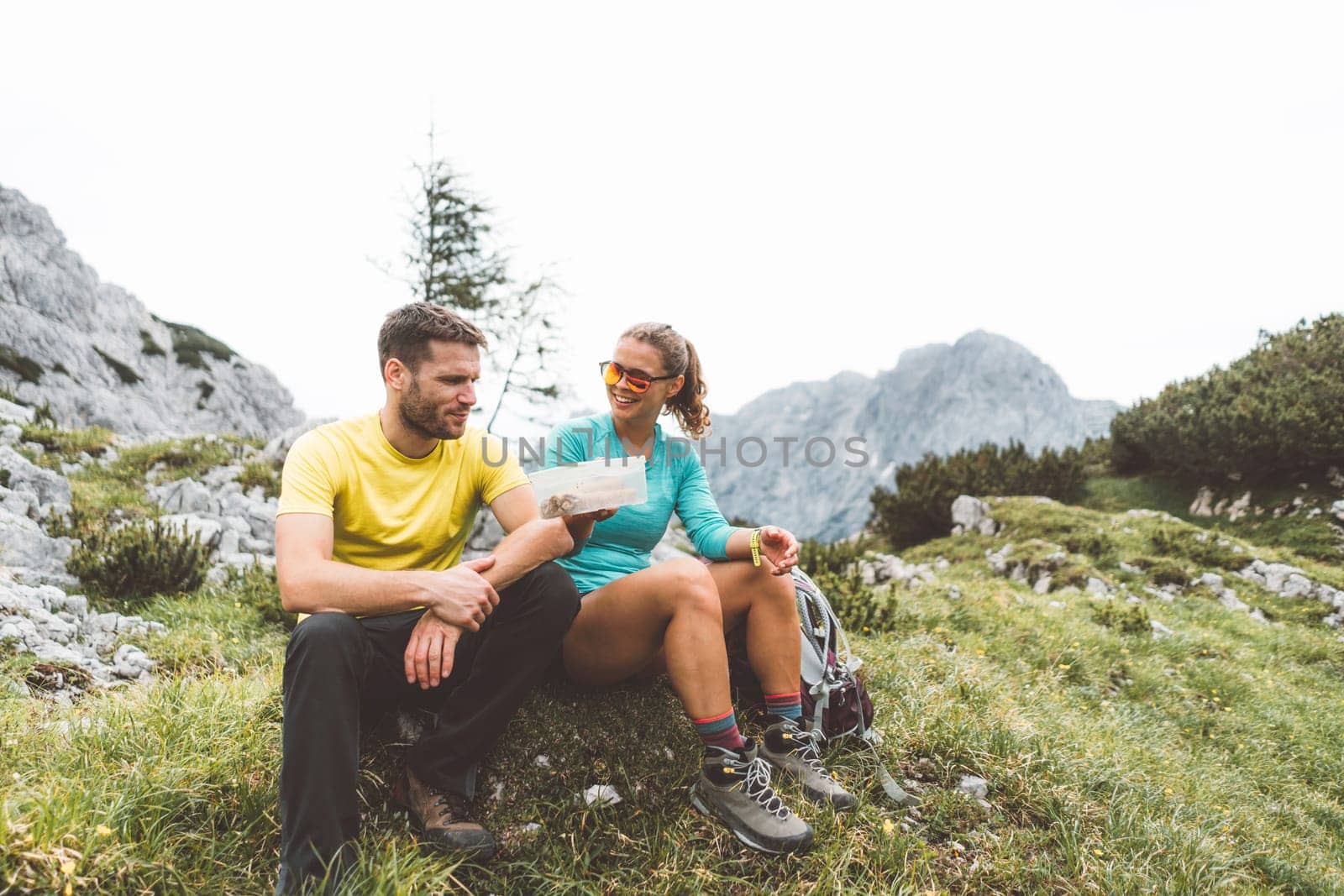 Couple of hikers sitting down on a grass field up in the mountains taking a stop for lunch break by VisualProductions