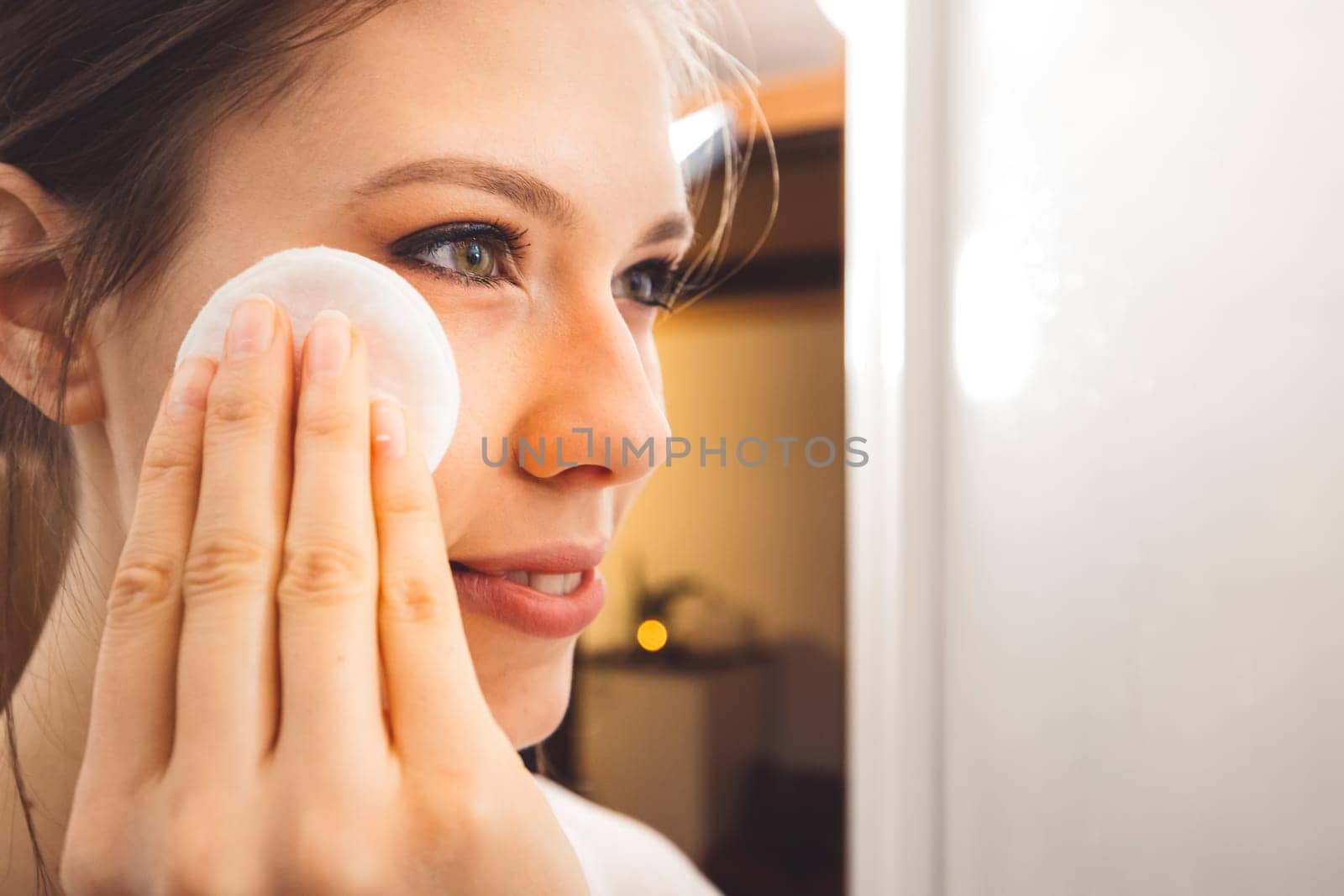 Close up side view of young woman removing make up off her face using a cotton pad and micellar water by VisualProductions