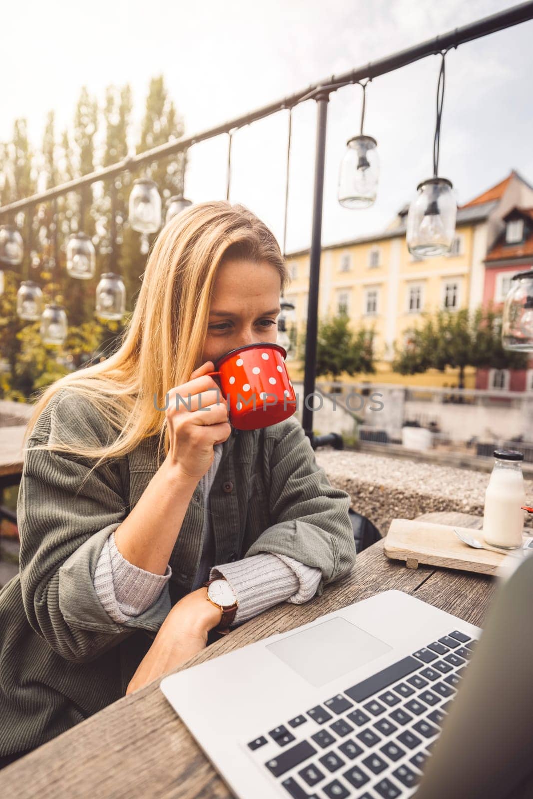 Vertical photo of a woman drinking coffee from a red cup while sitting outside at a cafe working on her laptop by VisualProductions
