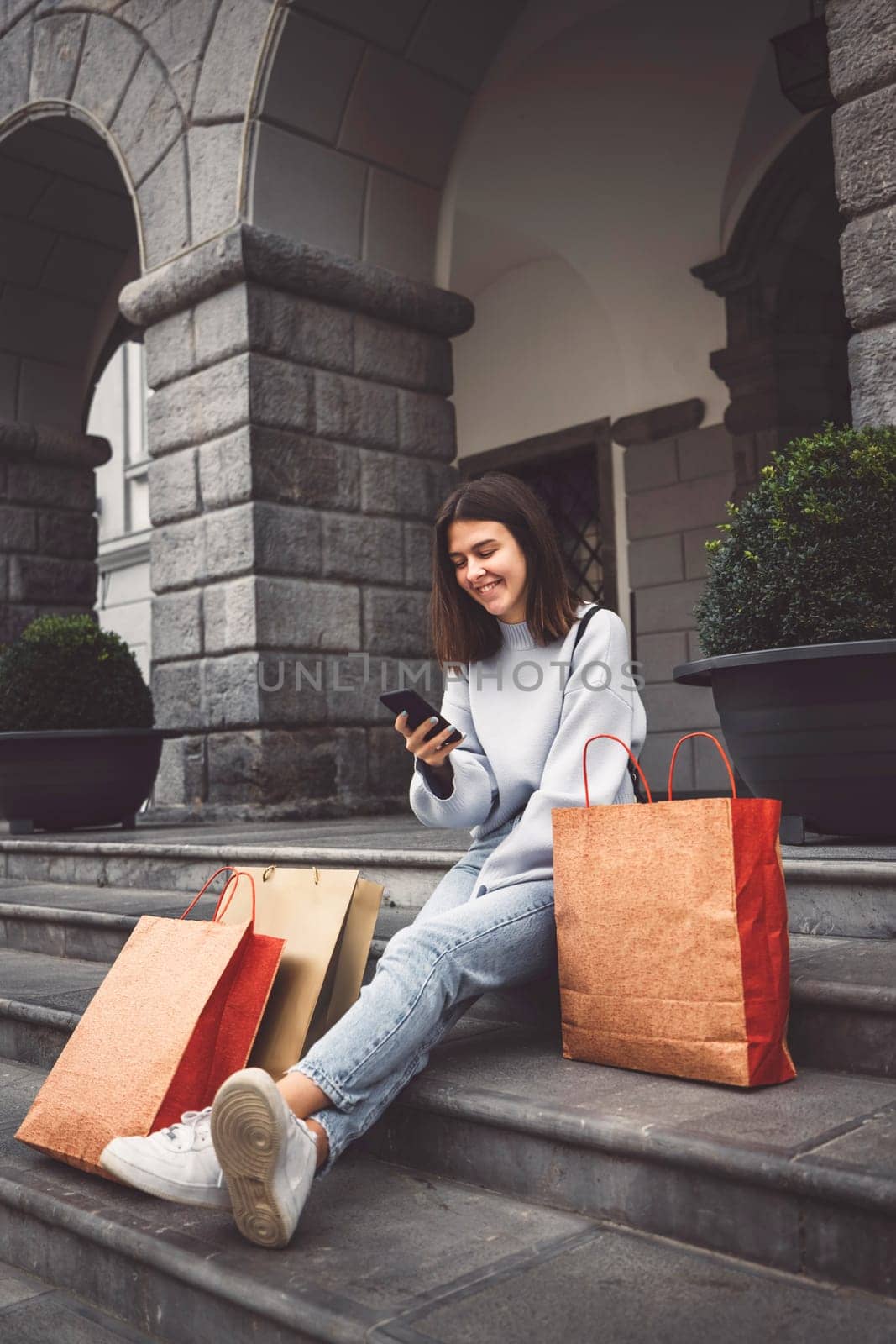 Vertical photo of brunette young woman sitting down on the stairs in the city with paper shopping bags around her by VisualProductions