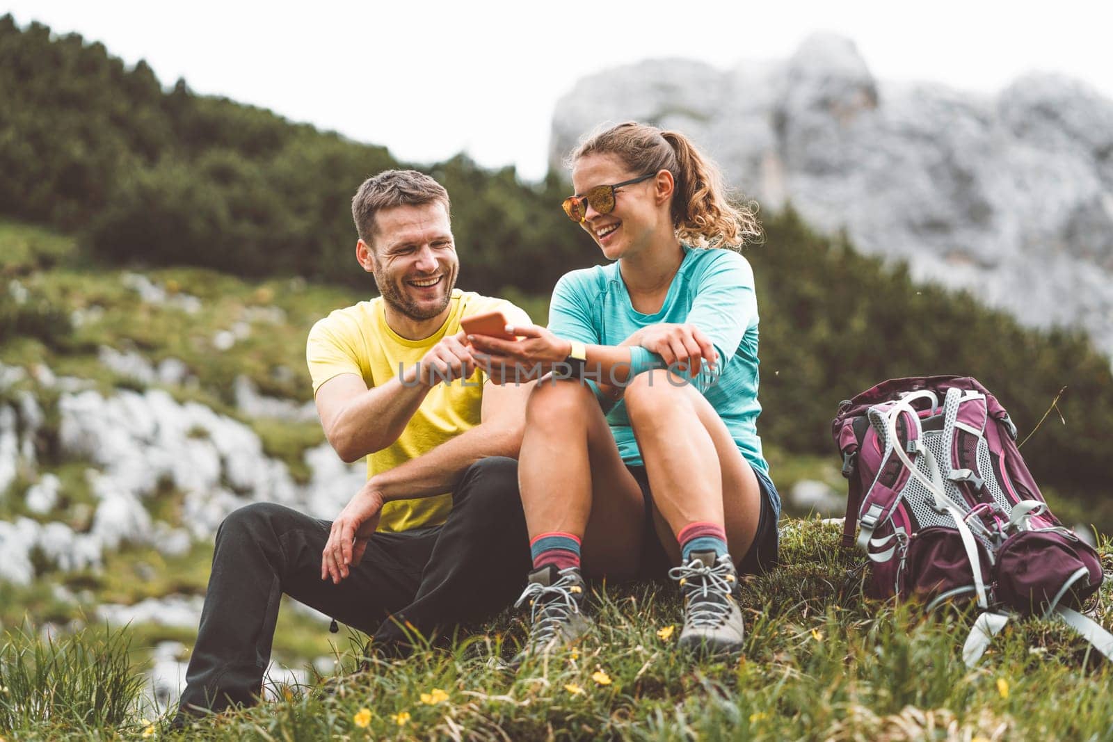 Smiling couple of hikers sitting down on the grass laughing while looking at the phone by VisualProductions